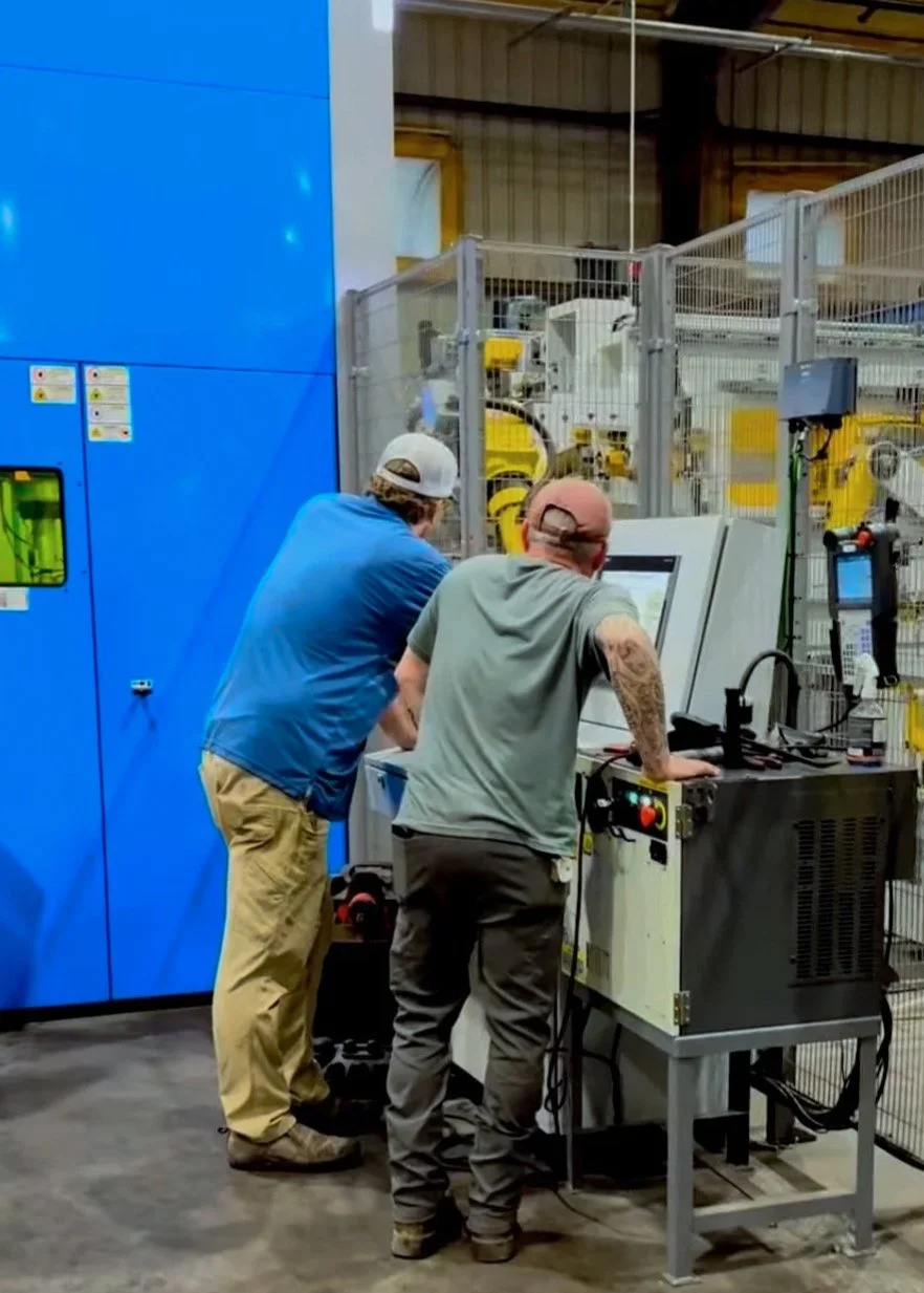 Two men working at a control panel in an industrial manufacturing facility, with machinery and safety fencing in the background.