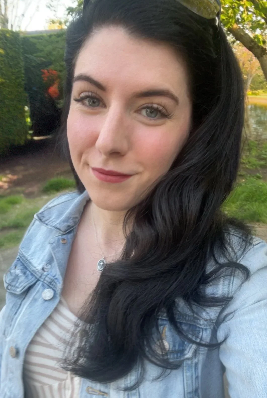 A young woman with long, wavy black hair and light skin smiling outdoors. She's wearing a denim jacket, a striped top, and a necklace. The background includes greenery and a body of water.