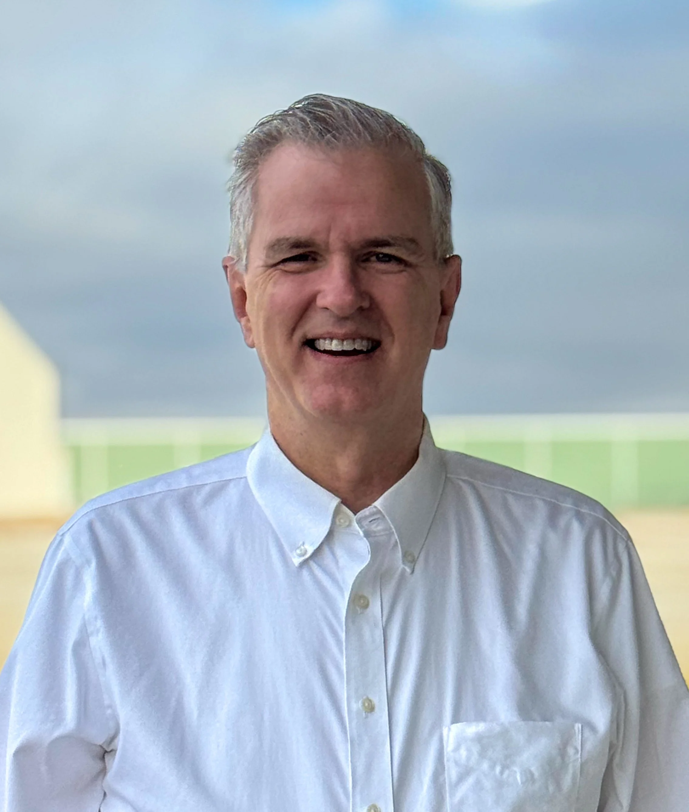 A middle-aged man with gray hair wearing a white button-up shirt, smiling outdoors with a partly cloudy sky and a faint landscape in the background.