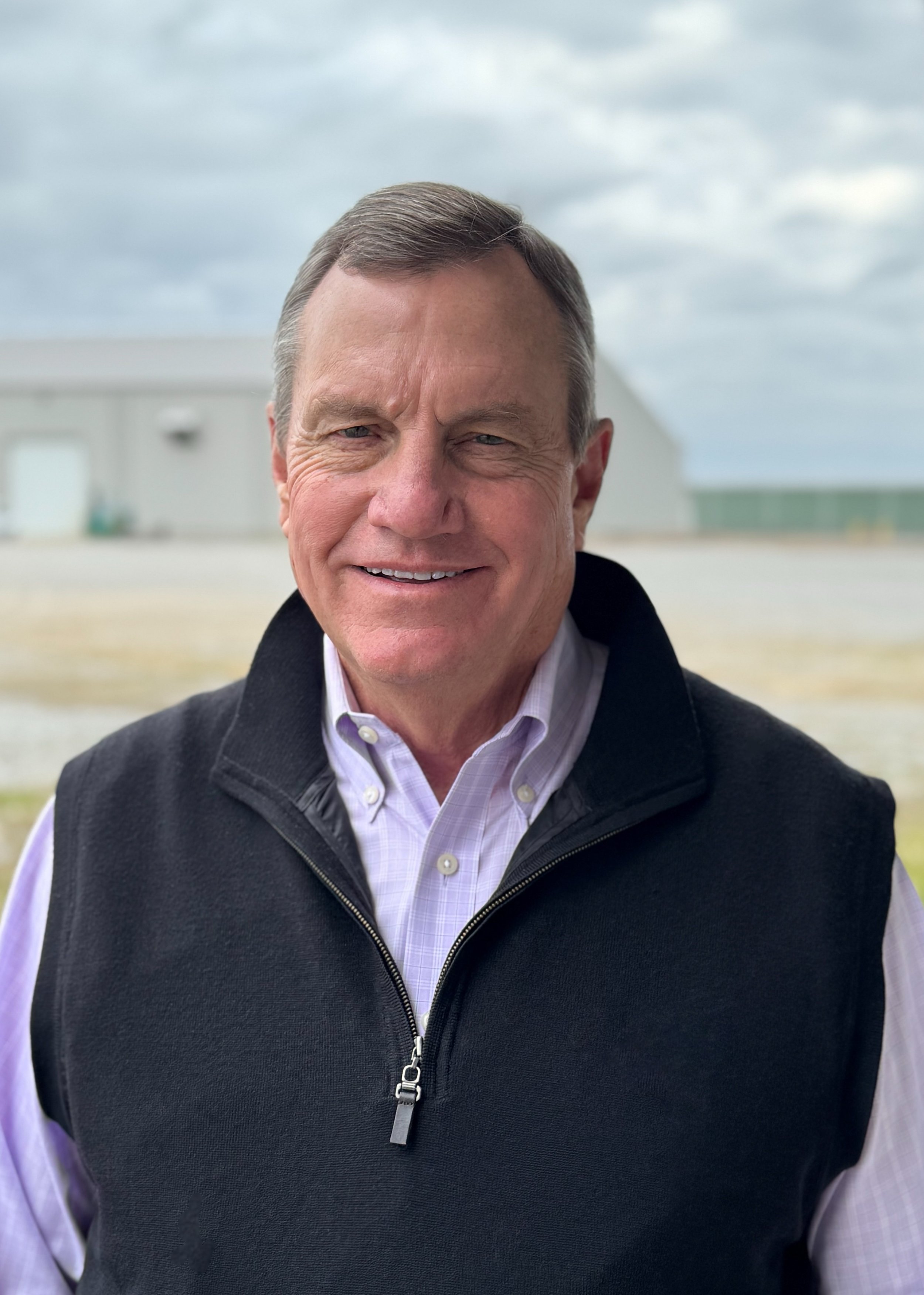 A smiling middle-aged man wearing a light purple collared shirt and a black vest outside on a cloudy day.