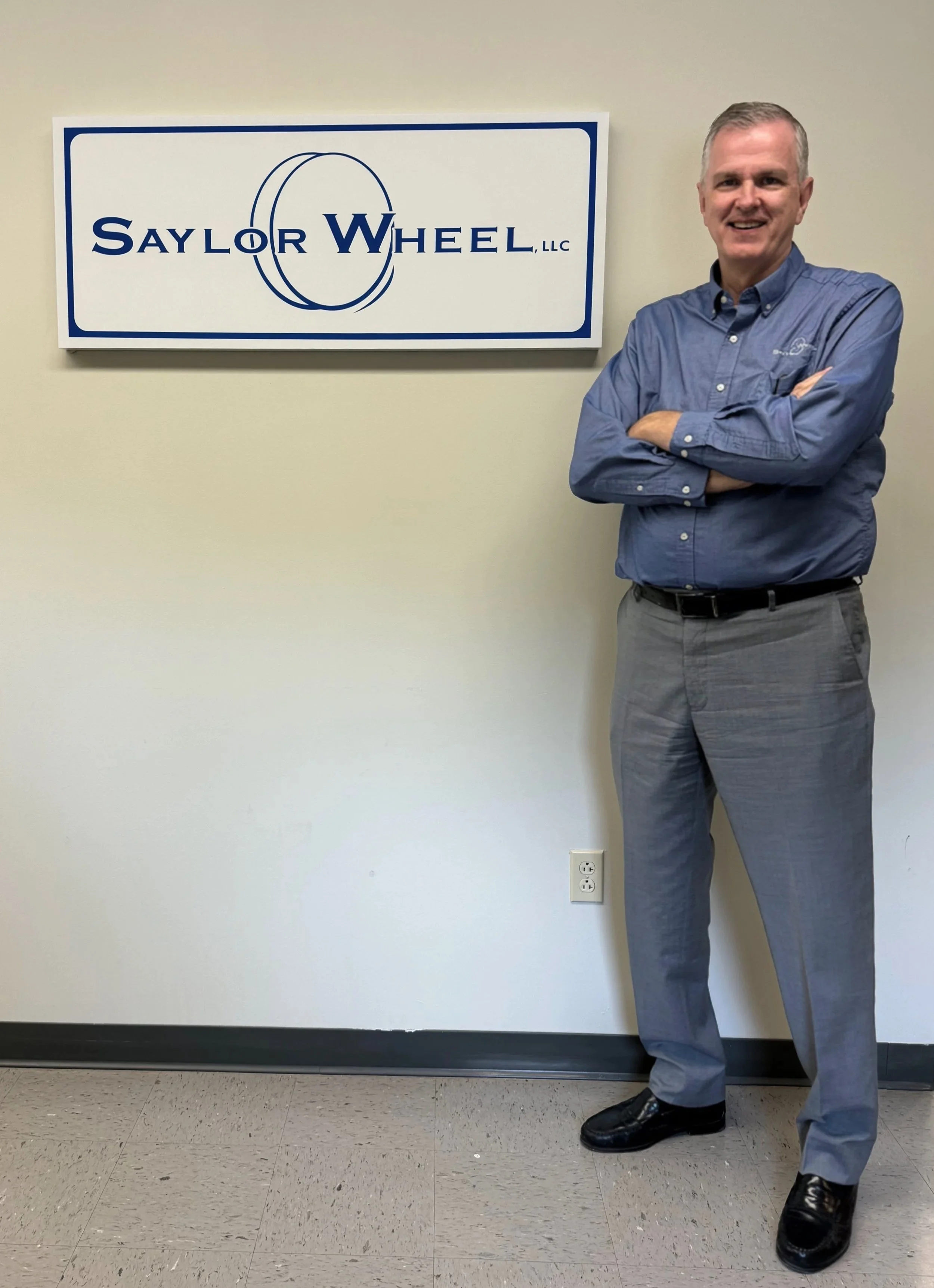 Image of Saylor wheel’s founder, Dave Saylor, in front of a Saylor Wheel sign with the blue logo