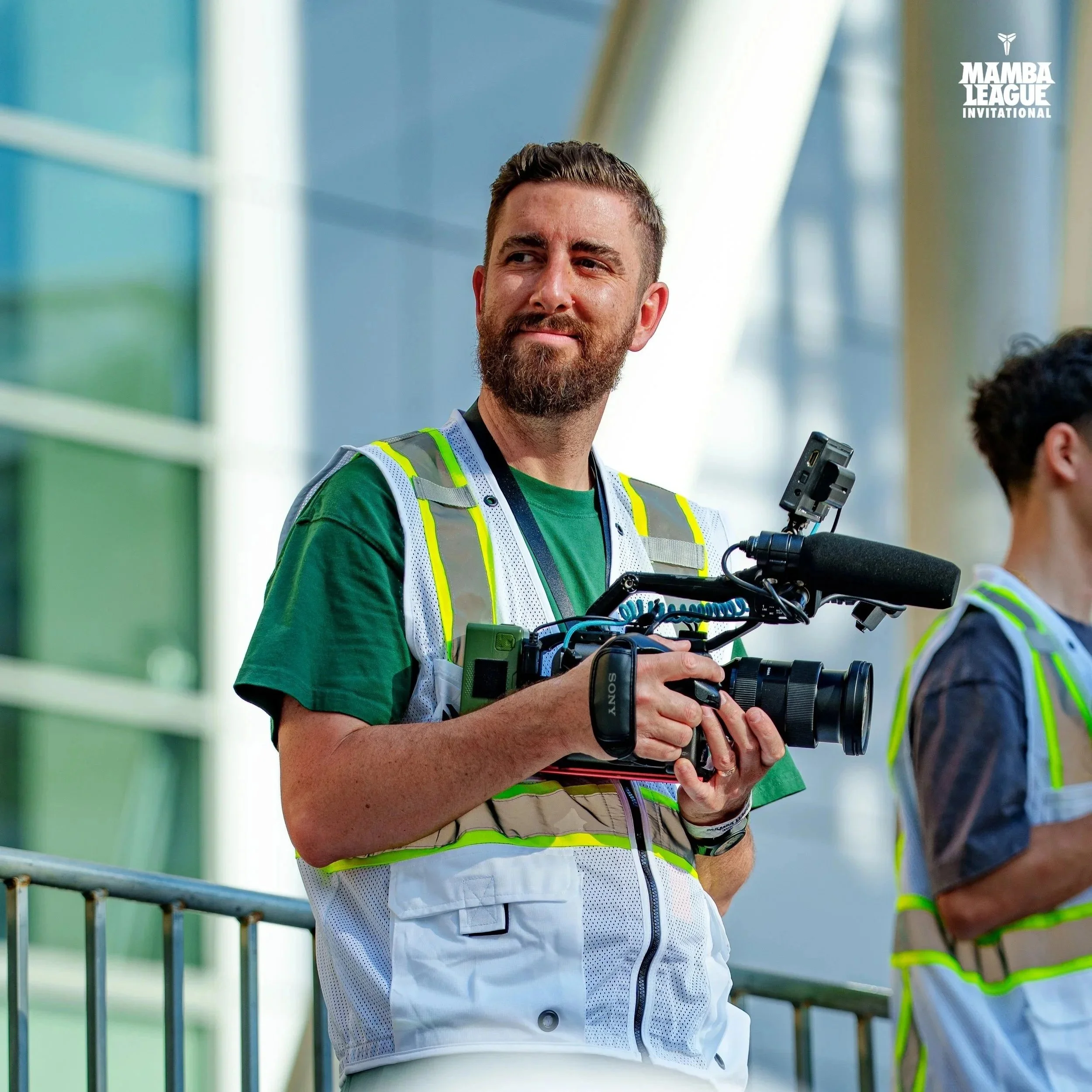 Man wearing a green shirt and reflective safety vest holding a professional video camera, standing outdoors near a modern glass building.