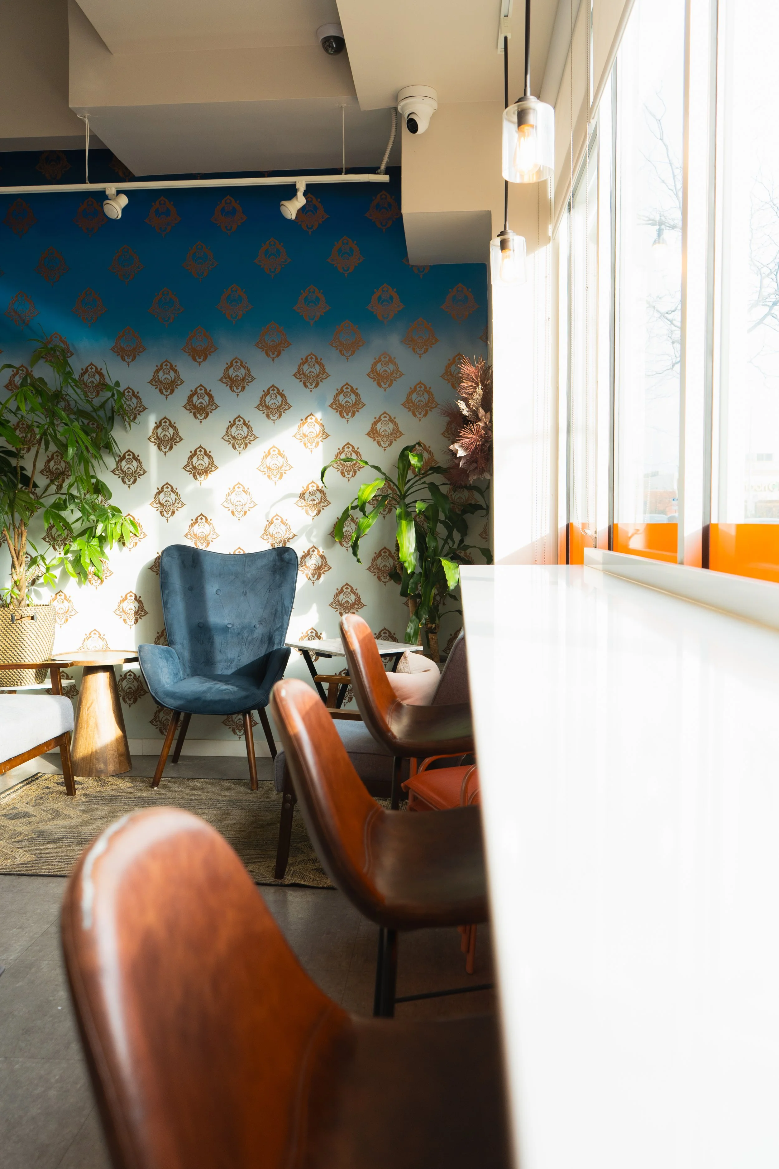 a long shot of a clean high bar with tall barstools. a comfy blue chair is in the background nestled among many plants.