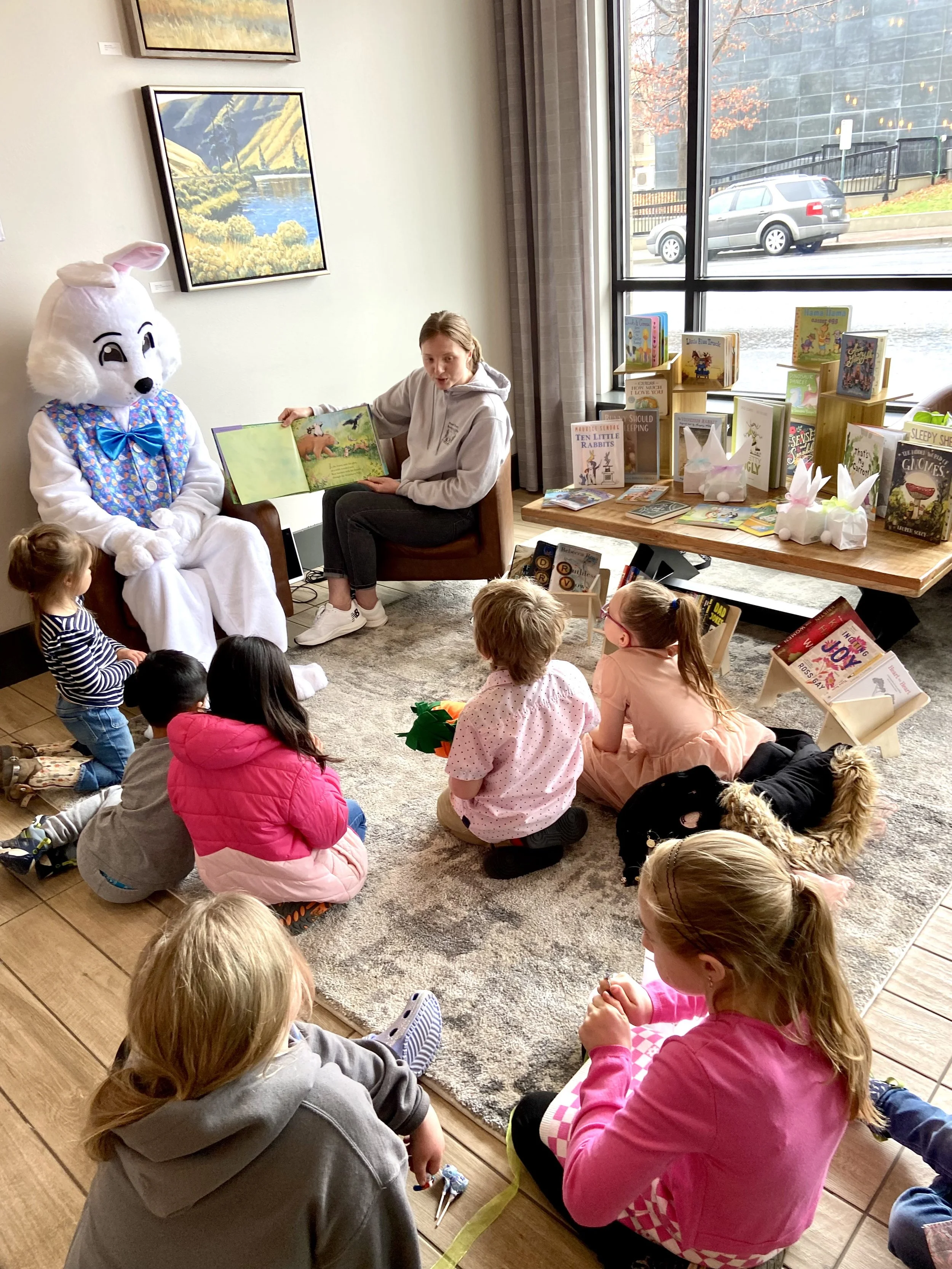 Children gathered for a storytime session with a person in an Easter bunny costume and another person reading a book. The setting is indoors with books displayed on a table and artwork on the walls.