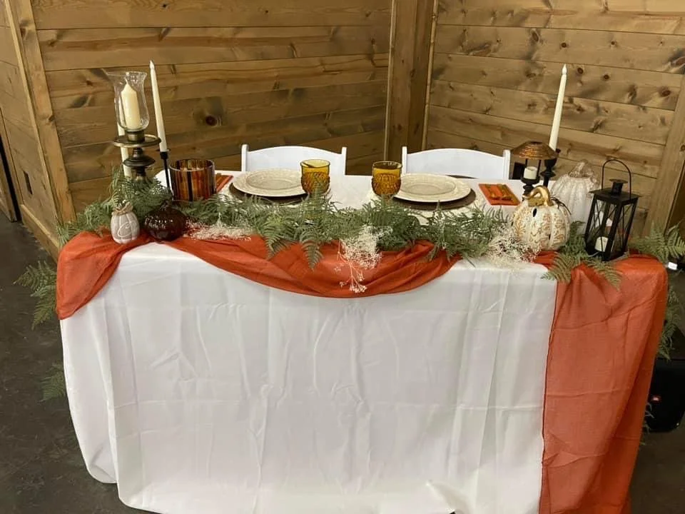 Decorated dining table with a white tablecloth and orange runner, adorned with greenery, pumpkins, candles, and lanterns, set against a wooden wall.