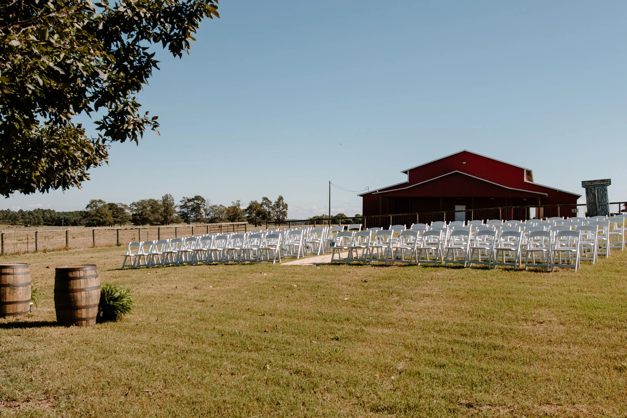 Outdoor wedding ceremony setup with white chairs arranged in rows facing a red barn on a sunny day, surrounded by fields and trees.
