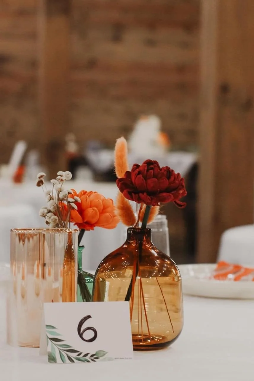 Table centerpiece with red and orange flowers in glass vases, table number 6 card, and blurred background.