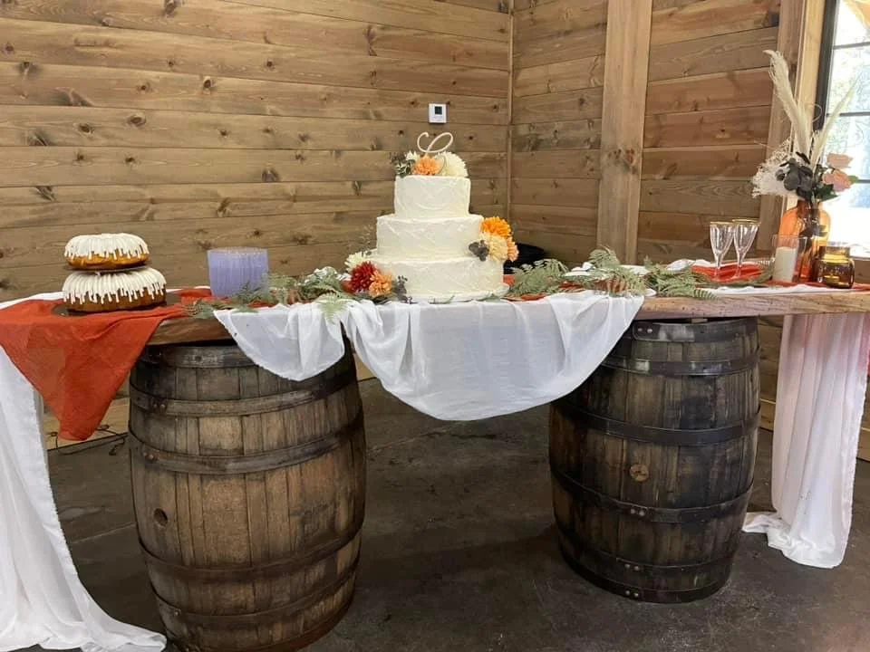 Rustic wedding cake table setup with a three-tiered cake decorated with flowers, positioned on a wooden barrel table. Additional cakes and decorations present, with a wooden backdrop.