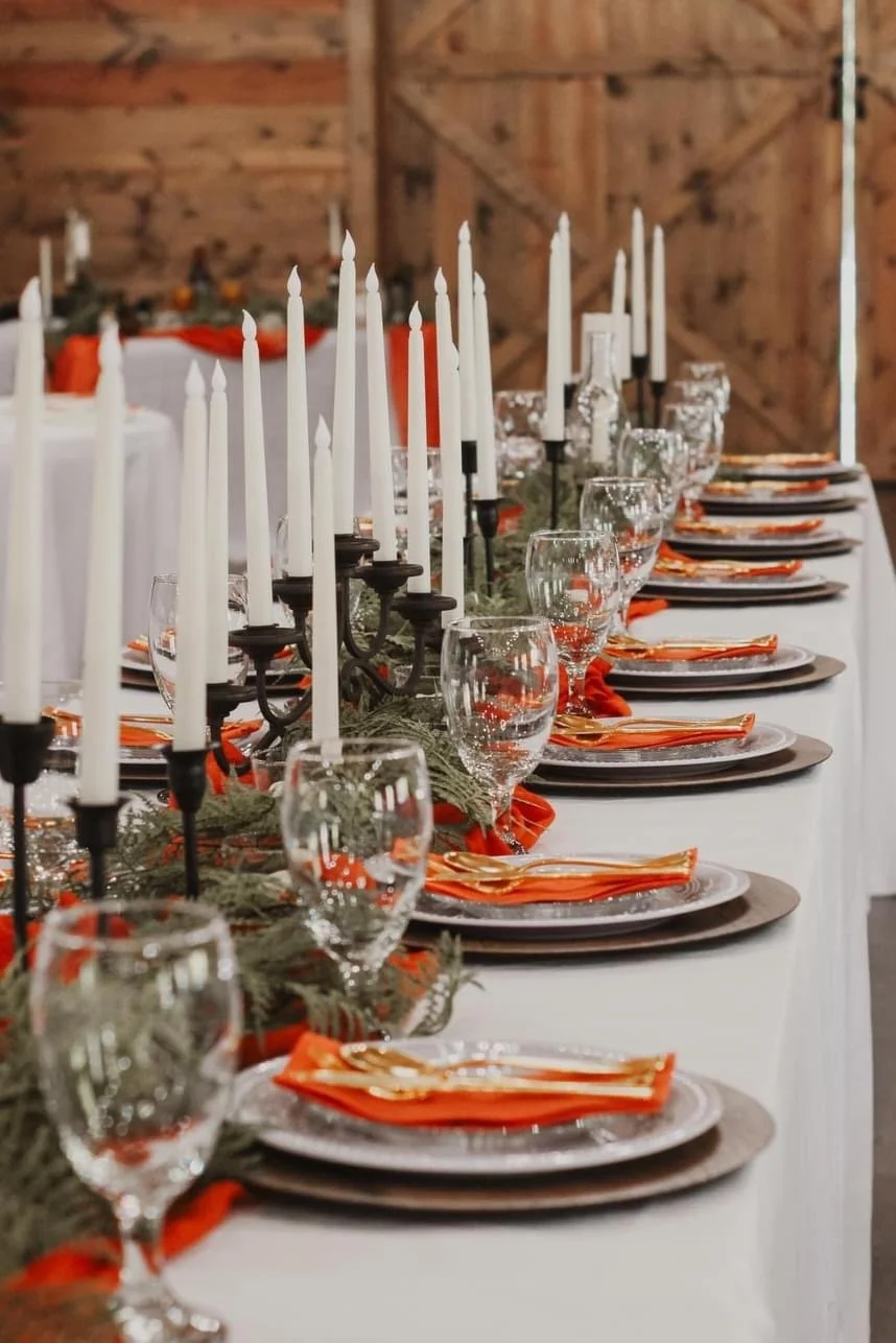 Elegant table setting with white candles, red napkins, glassware, plates, and greenery decoration in a rustic venue.