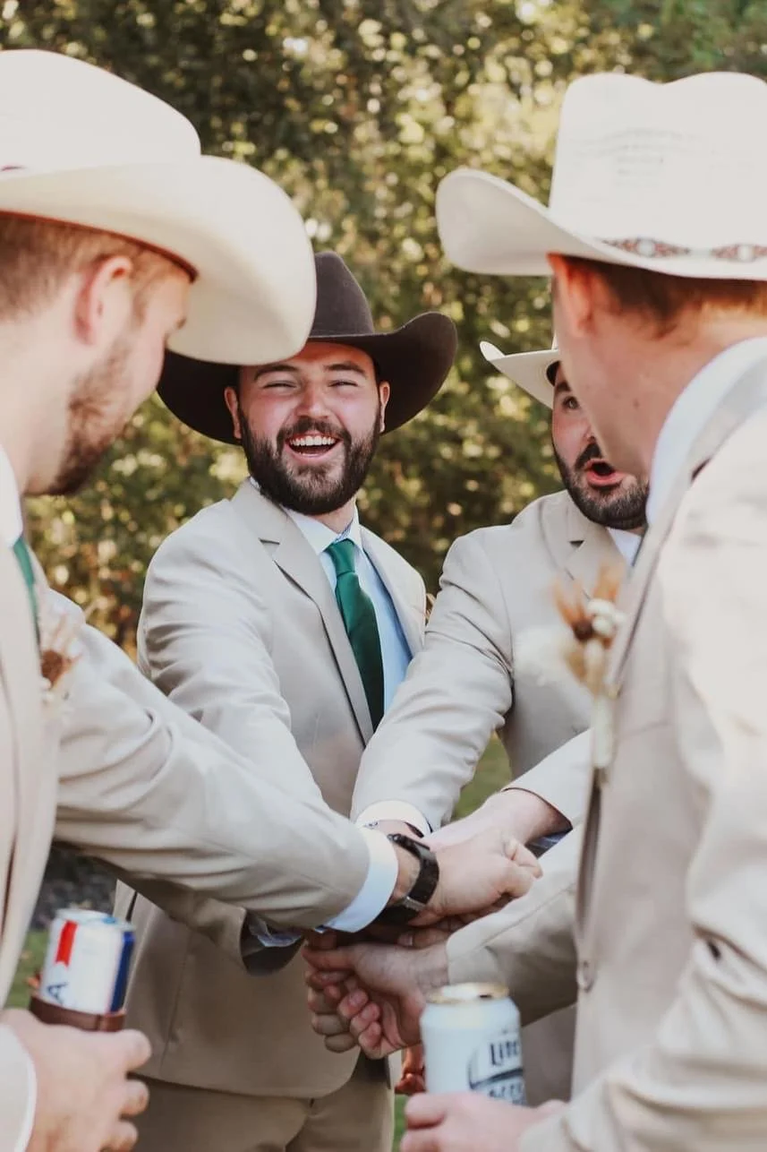 Group of men in suits and cowboy hats, hands together in a gesture of unity, outdoors.