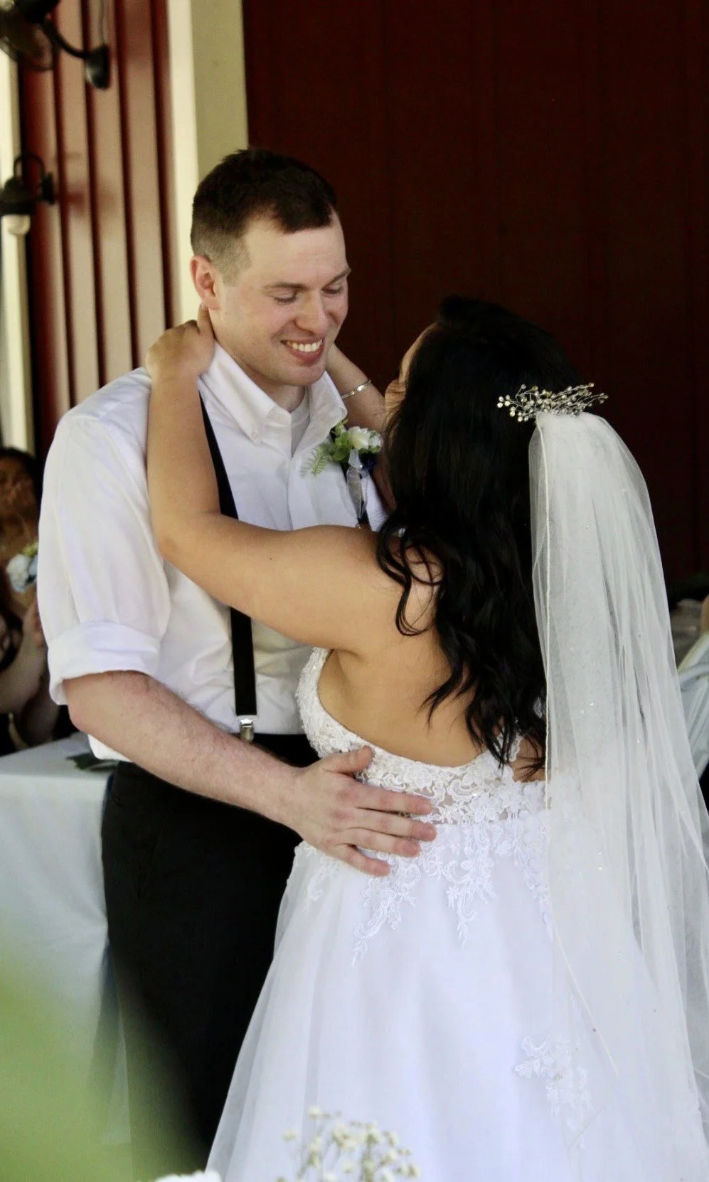 Bride and groom embracing during wedding dance, bride wearing a veil and white dress, groom in a white shirt with suspenders.