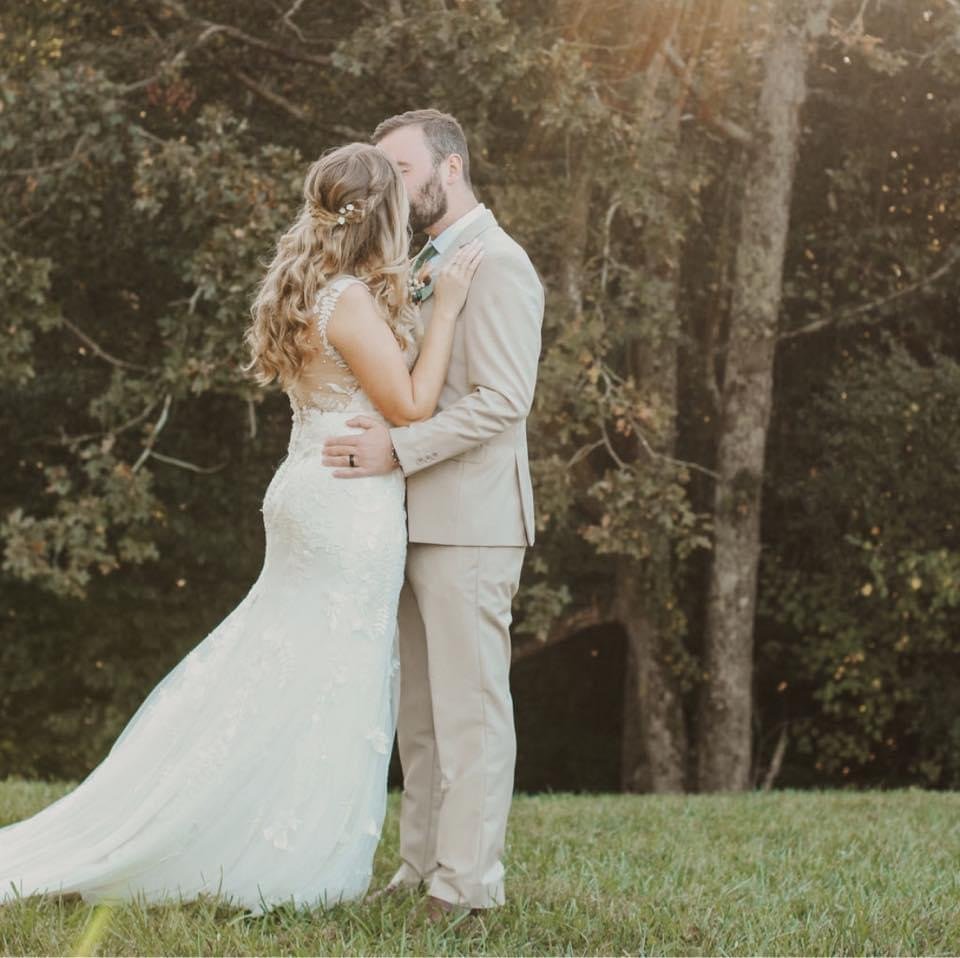 A bride and groom kissing outdoors in a park, surrounded by trees and grass.
