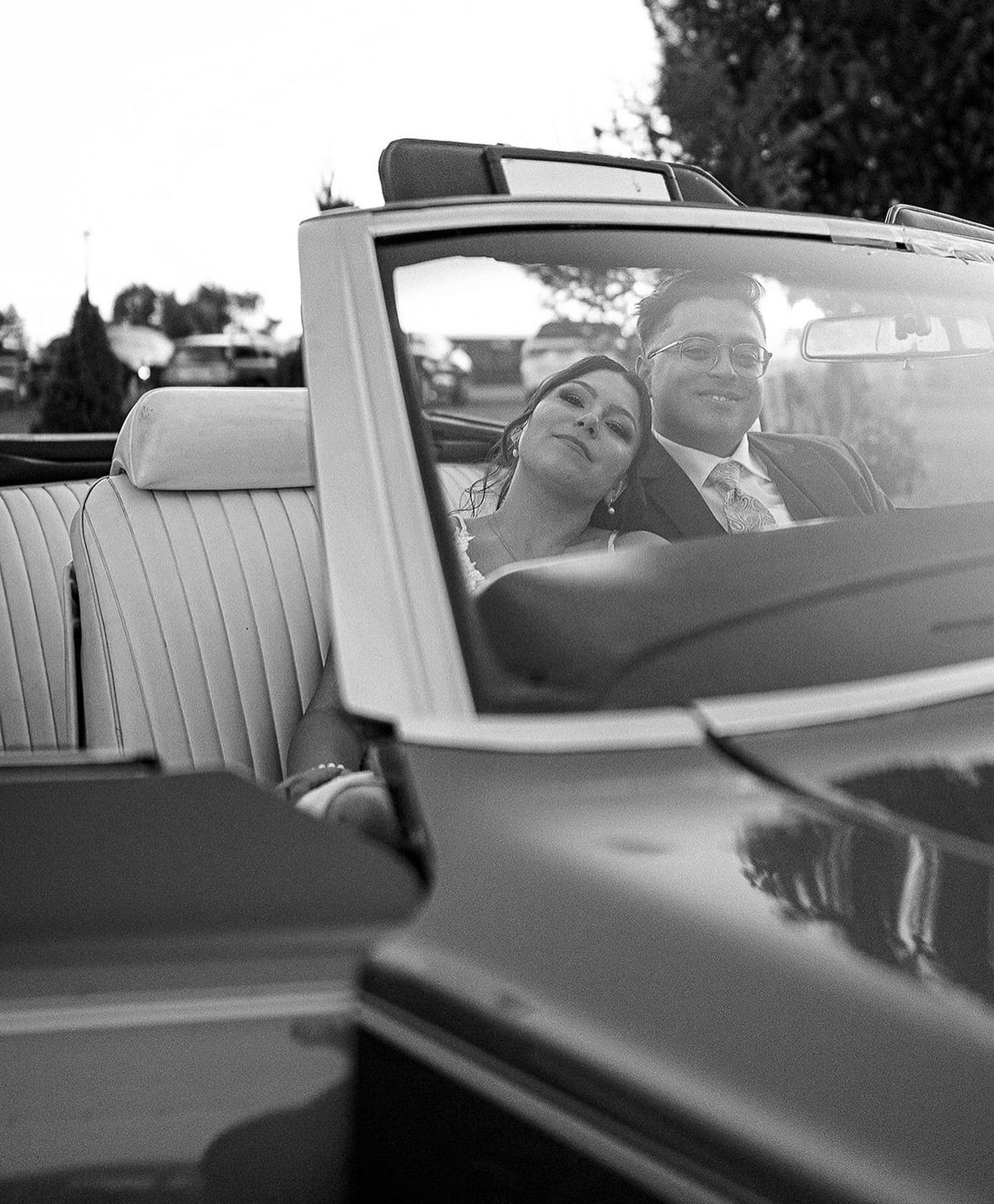 Black and white photo of a couple sitting in the front seats of a convertible car, appearing happy and relaxed, with trees and other vehicles in the background.