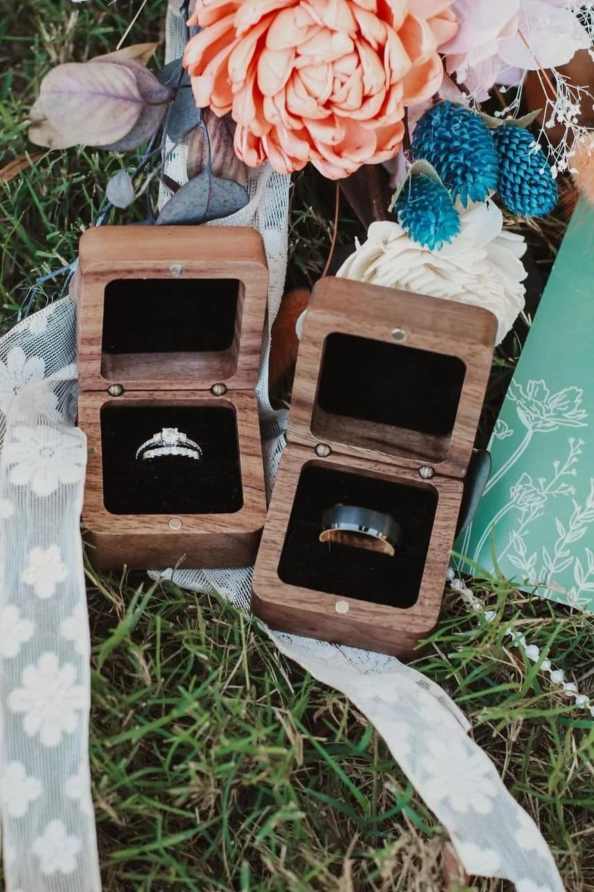Two open wooden ring boxes on grass with lace and flowers, containing a silver ring and a dark band.