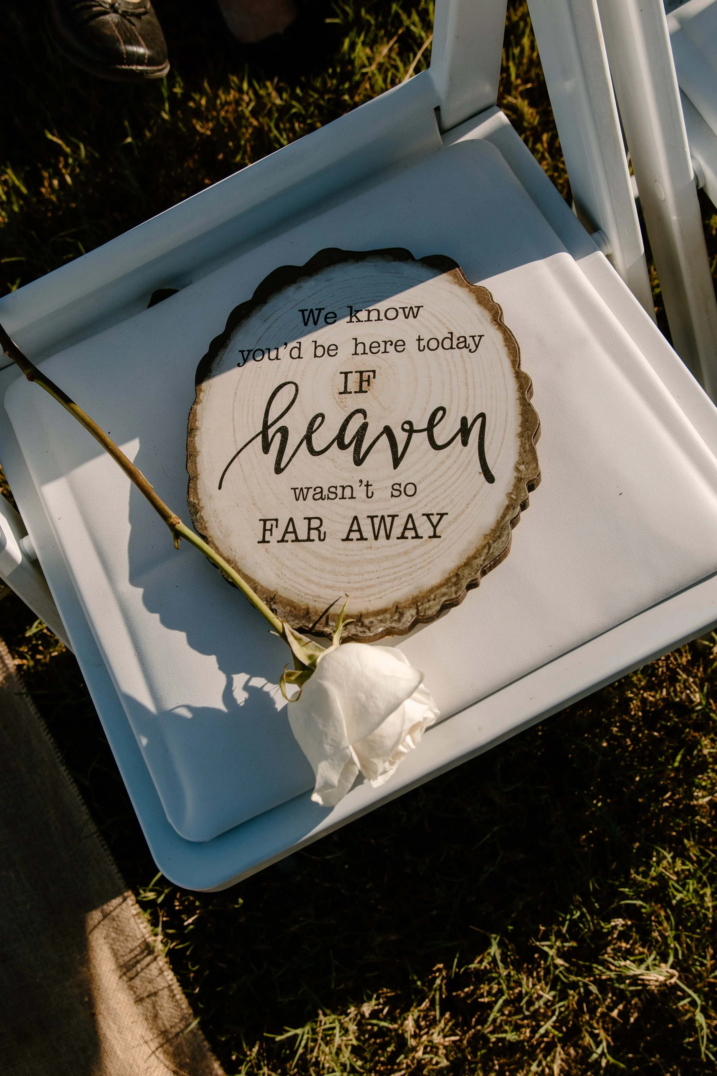 Memorial sign on chair with text "We know you'd be here today if heaven wasn't so far away" and a white rose.