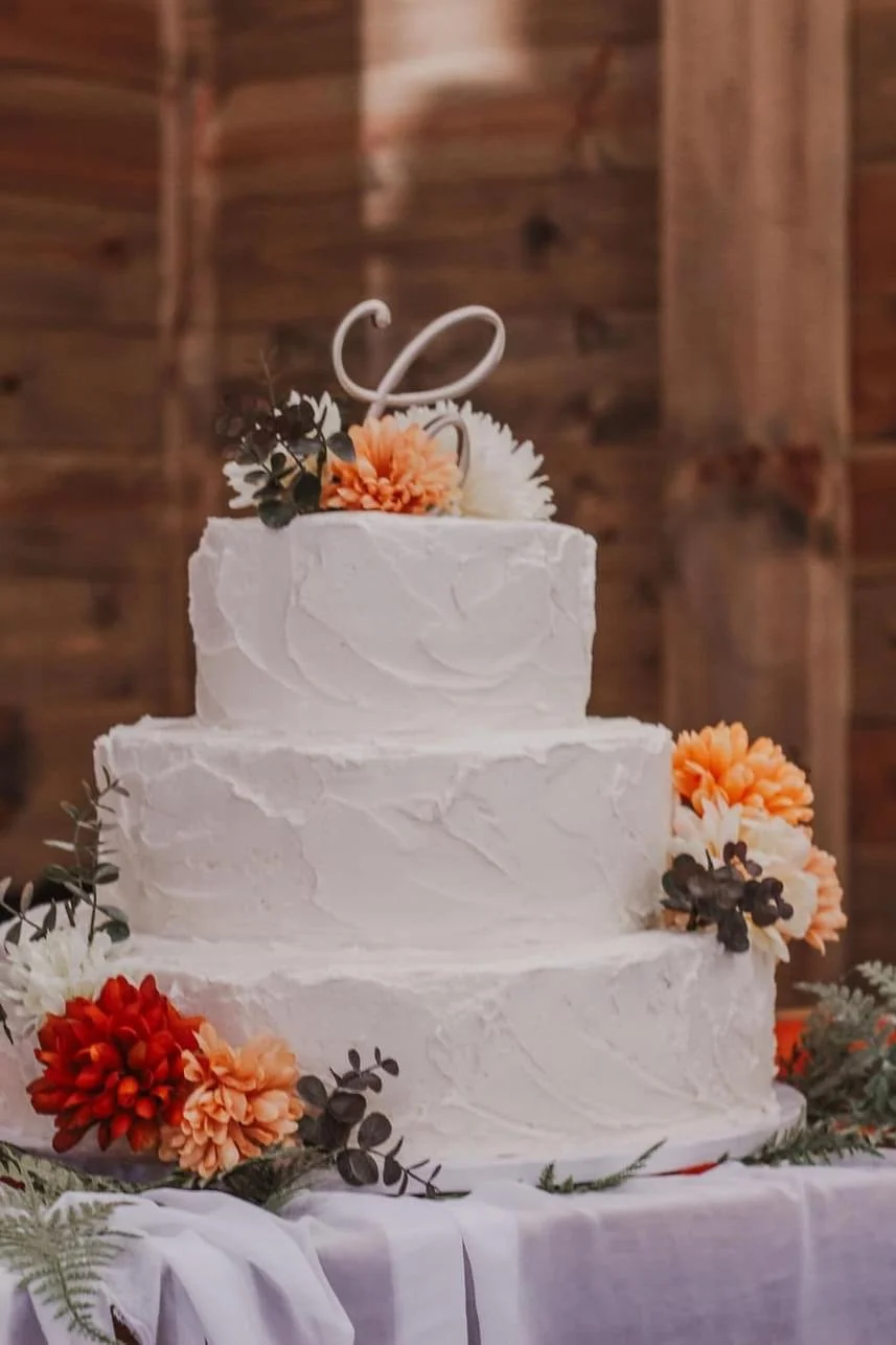 Three-tier white wedding cake with floral decorations and a wire topper on a draped table.