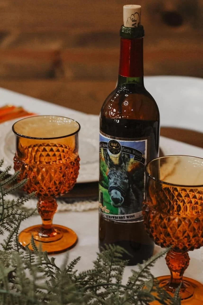 Bottle of strawberry wine with a donkey label, between two ornate amber goblets, on a table with plant decoration.