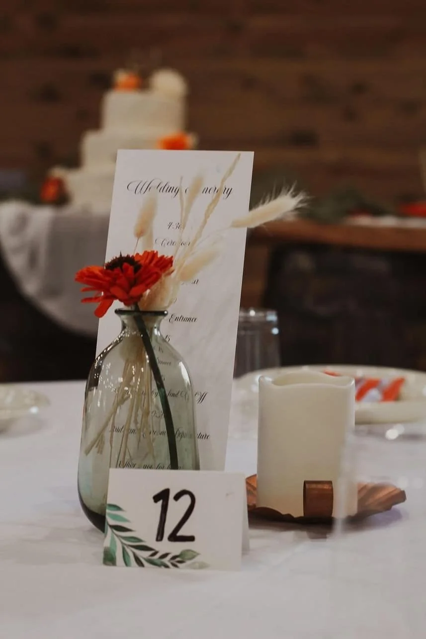 Table setup with a vase holding red flowers and pampas grass, a wedding ceremony program, and a table number card labeled '12'. A lit candle sits beside them with a blurred background featuring a tiered cake.