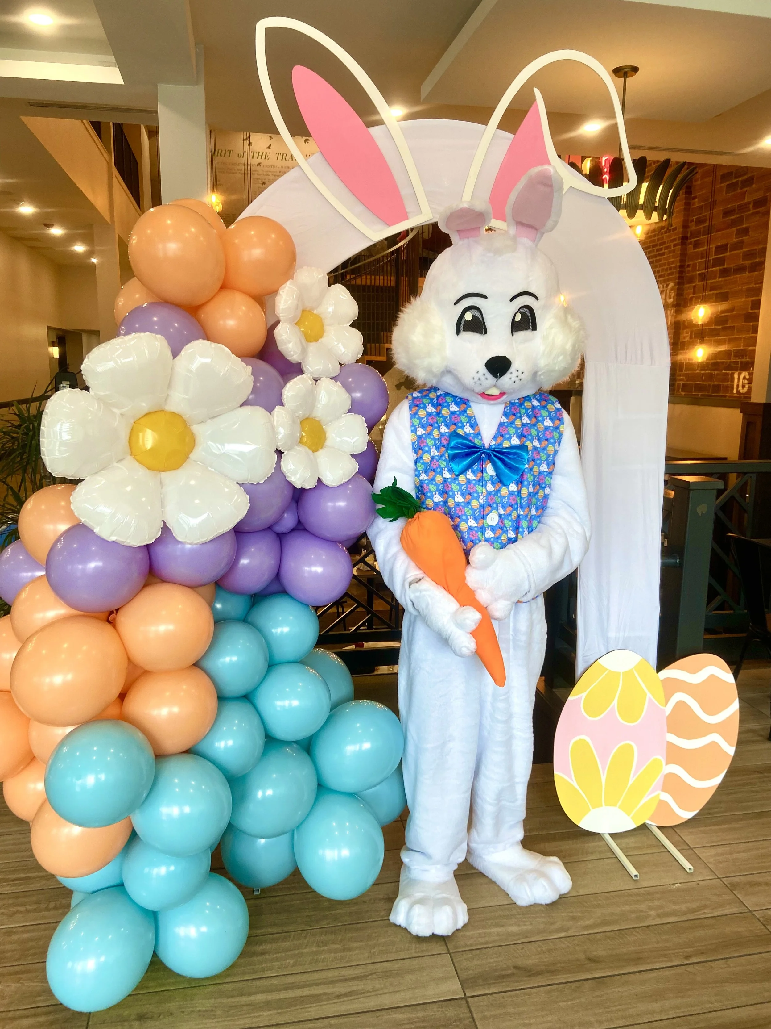 Easter bunny costume character holding a carrot, standing next to a colorful balloon arch and Easter decorations with flowers and eggs, in an indoor setting.