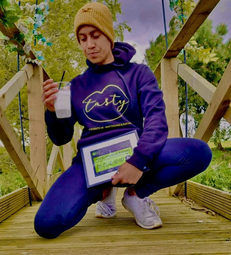 A person crouching on a wooden platform outdoors, holding a drink and a tablet with a food hygiene rating. They are wearing a yellow knit beanie, a purple hoodie with 'Tasty' written on it, and white sneakers.