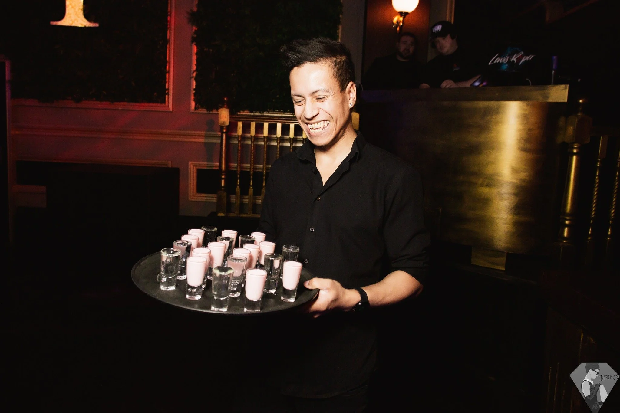 A smiling man in black shirt holding a tray with shot glasses and pink drinks in a dimly lit venue.