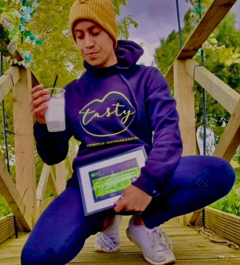 A young man crouching on a wooden deck holding a beverage in a plastic cup and a tablet with a colorful screen, wearing a purple hoodie, white sneakers, and a yellow beanie, outdoors surrounded by green trees.