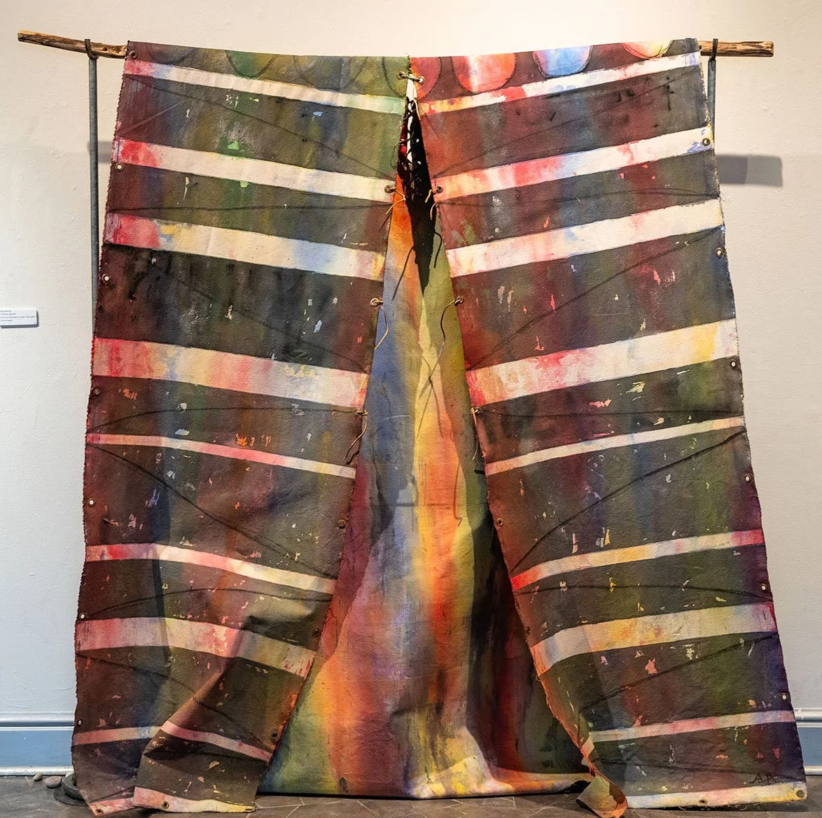 An art installation of layered, painted wooden planks hung from a wooden rod, creating a colorful abstract piece with dark and vibrant hues.