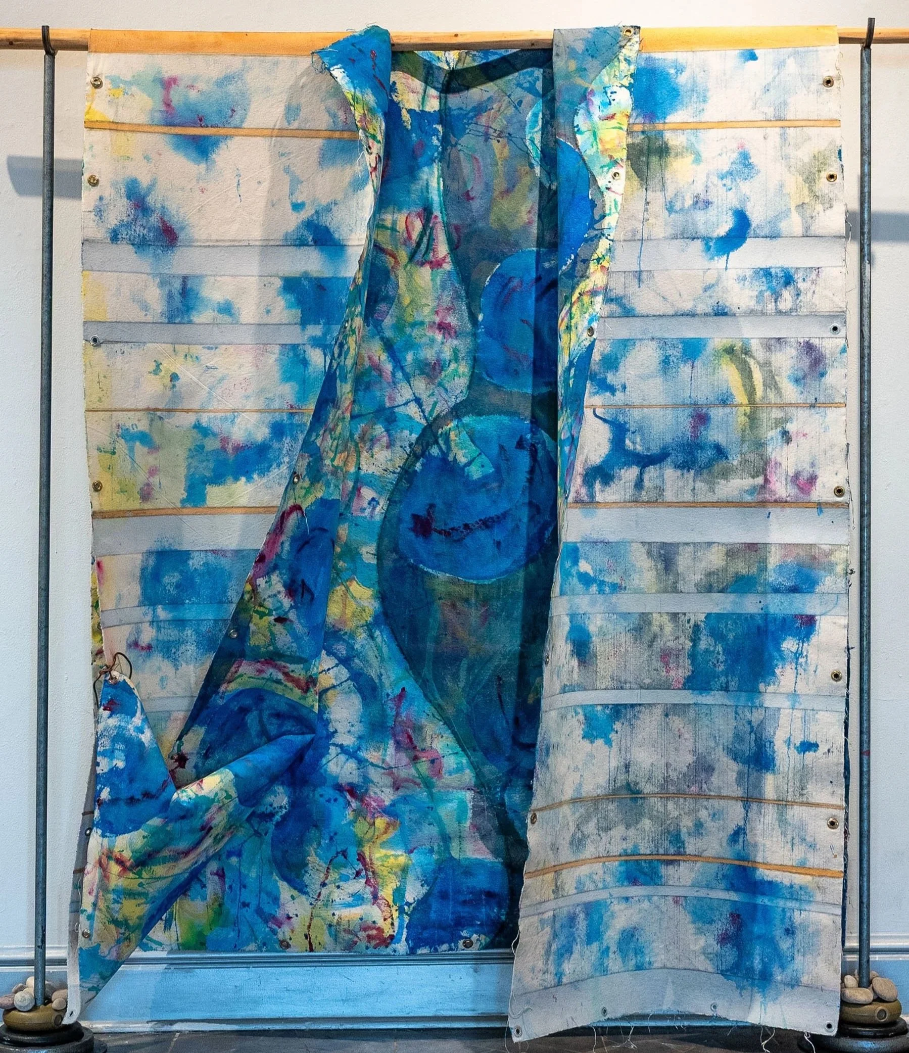 An abstract, multicolored fabric with blue, yellow, red, and green, draped over a wooden and metal frame with a painted wooden panel background. The fabric features swirling patterns and layered textures.