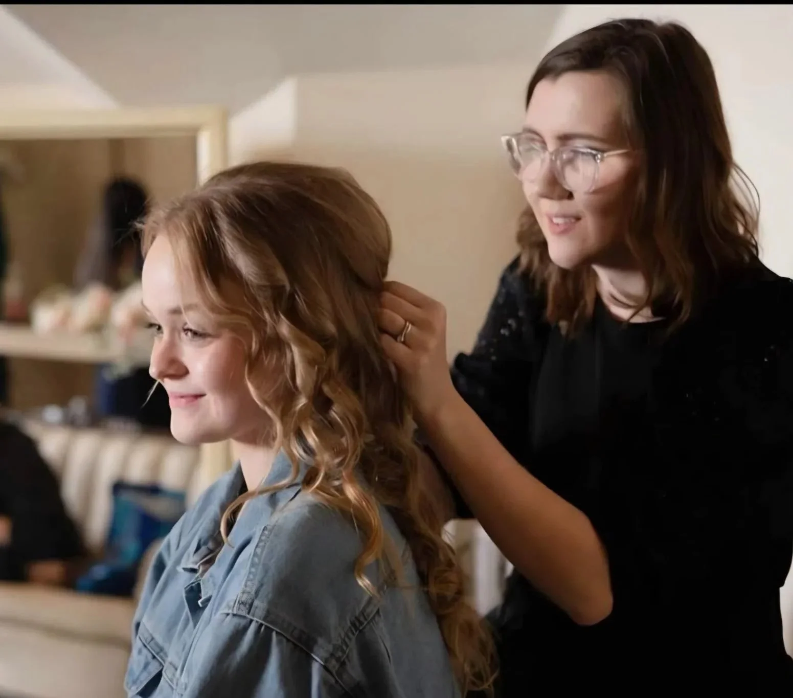 A woman with glasses styles a young woman’s curly red hair, both smiling, in a cozy room.
