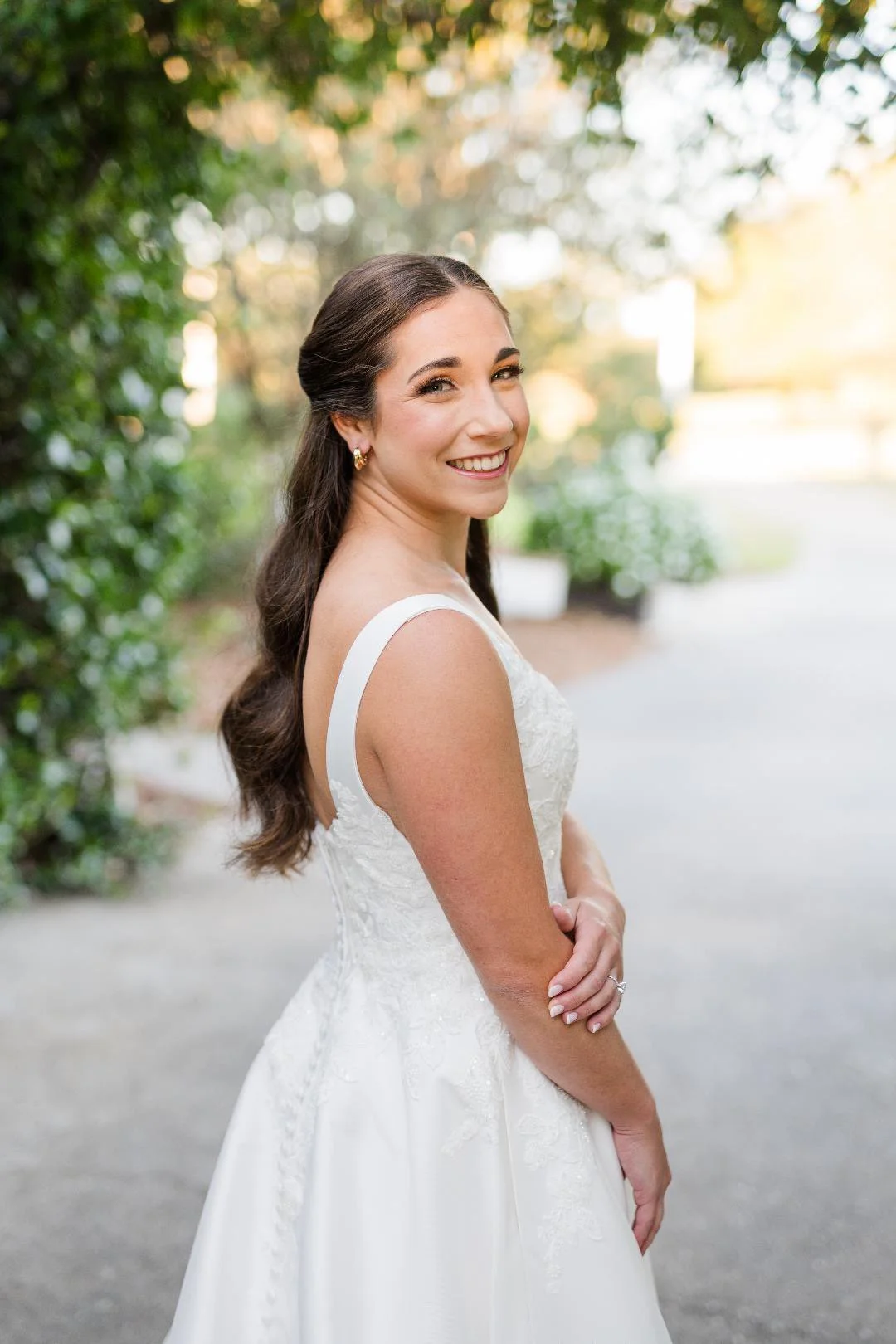 A smiling woman in a white wedding dress standing outdoors on a path surrounded by greenery with sunlight filtering through the trees in the background.