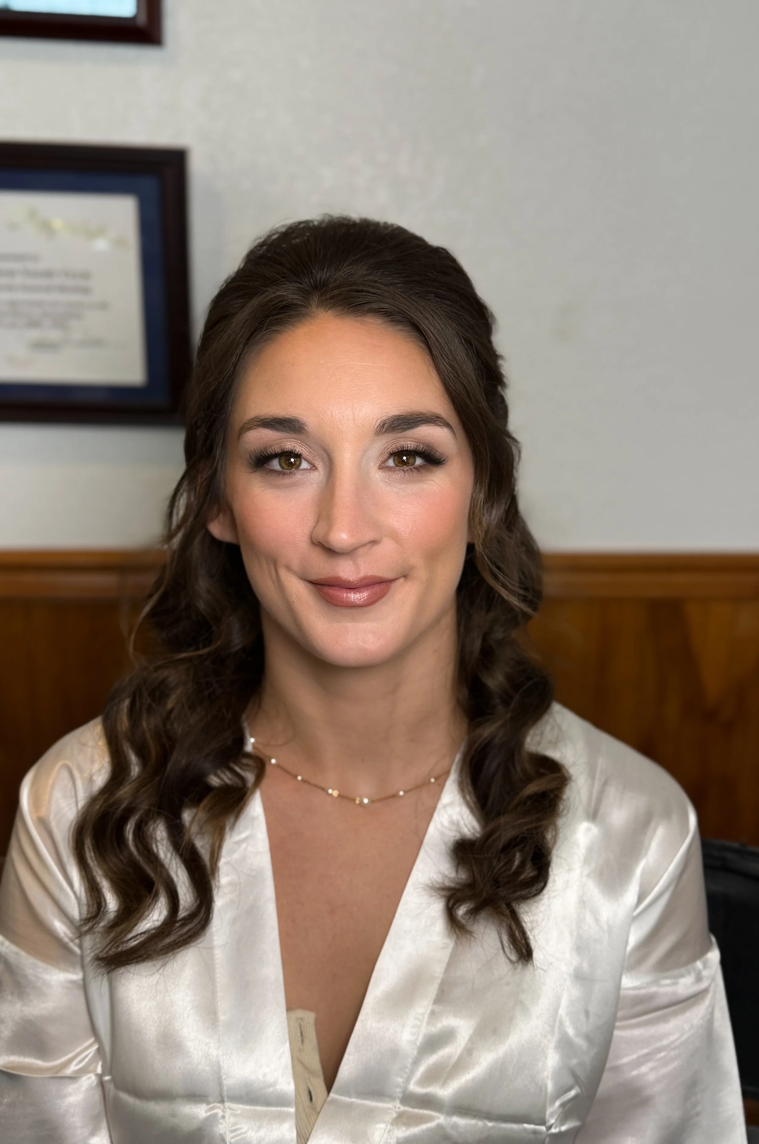 A woman with long brown curly hair, wearing a white satin top and a delicate necklace, smiling softly at the camera. There are framed diplomas or certificates on the wall behind her.