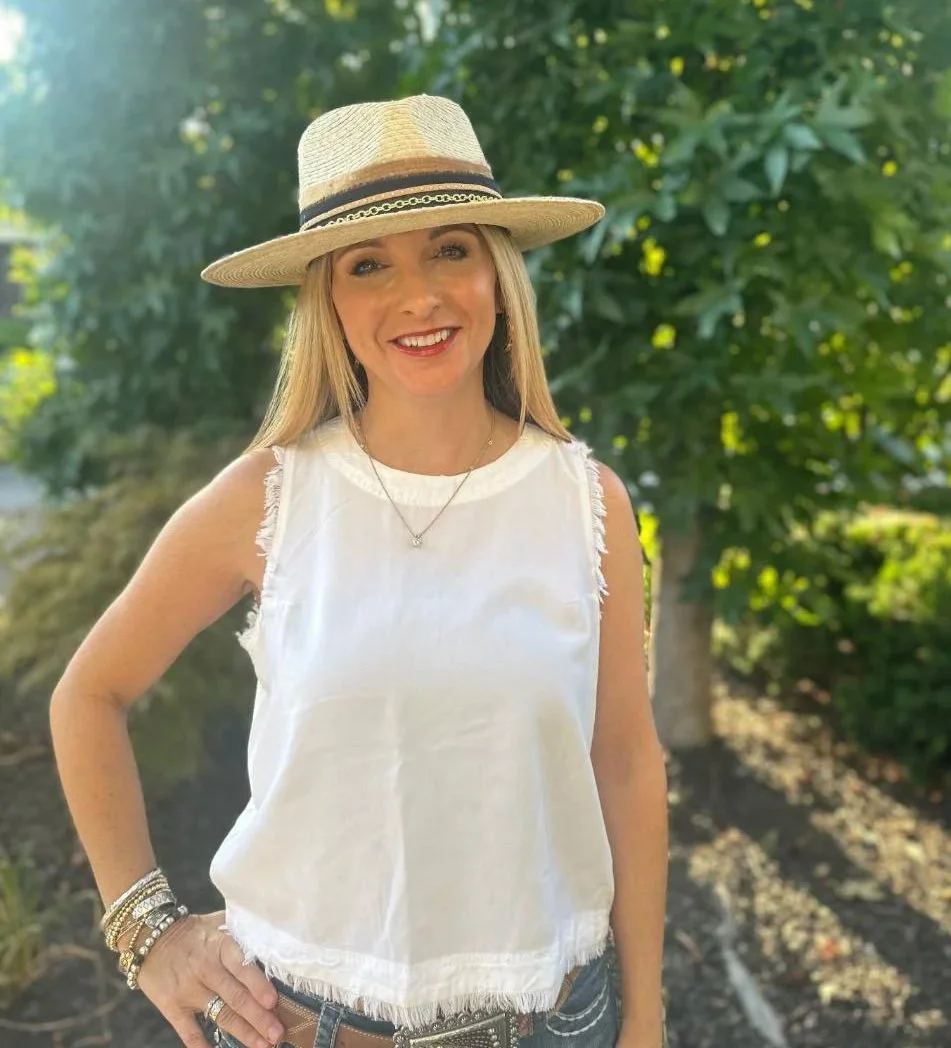 A smiling woman wearing a wide-brimmed straw hat, sleeveless white top with frayed edges, multiple bracelets, rings, and a necklace, standing outdoors with green trees in the background.