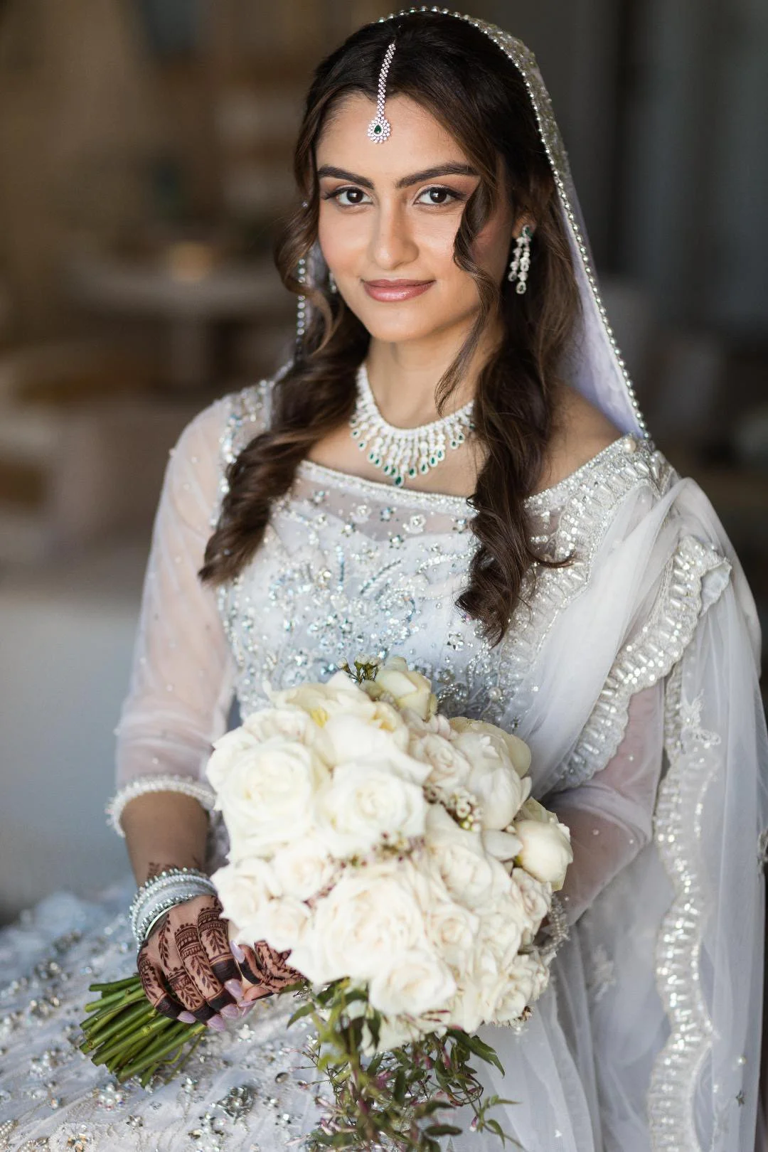 A bride in traditional white wedding attire holding a bouquet of white roses, adorned with jewelry including a necklace, earrings, and a headpiece, with henna designs on her hands and a slight smile.