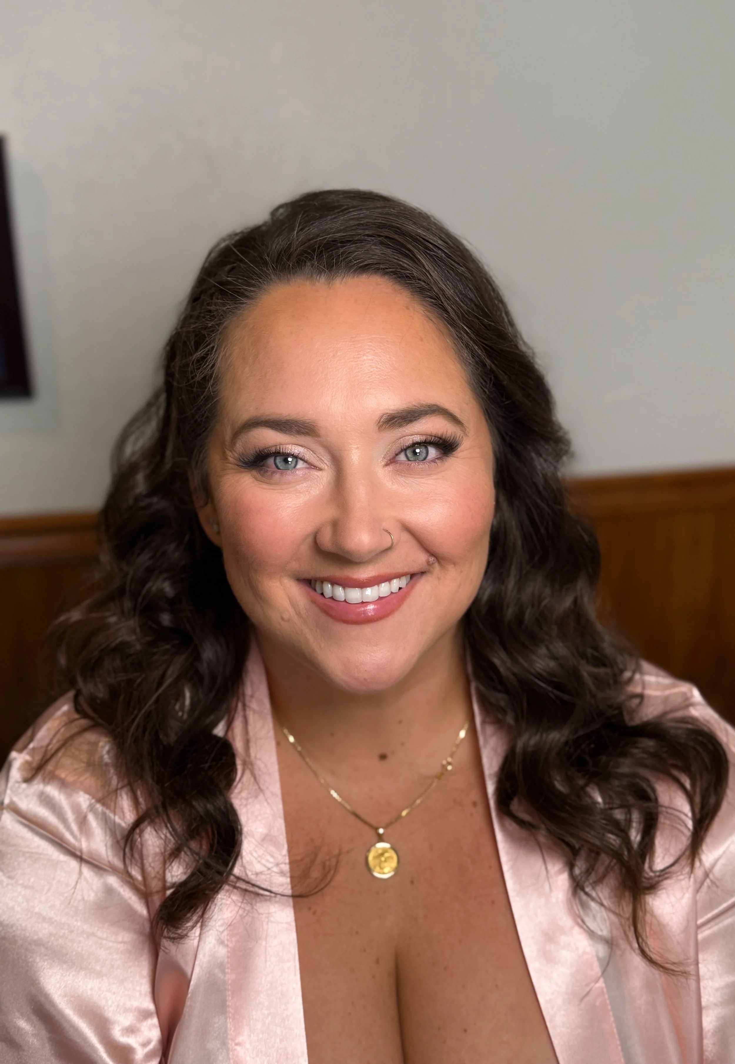 A woman with wavy brown hair, blue eyes, and wearing makeup, smiling at the camera, dressed in a light pink satin top with a gold necklace, sitting indoors with a wooden wall and a grayish background.