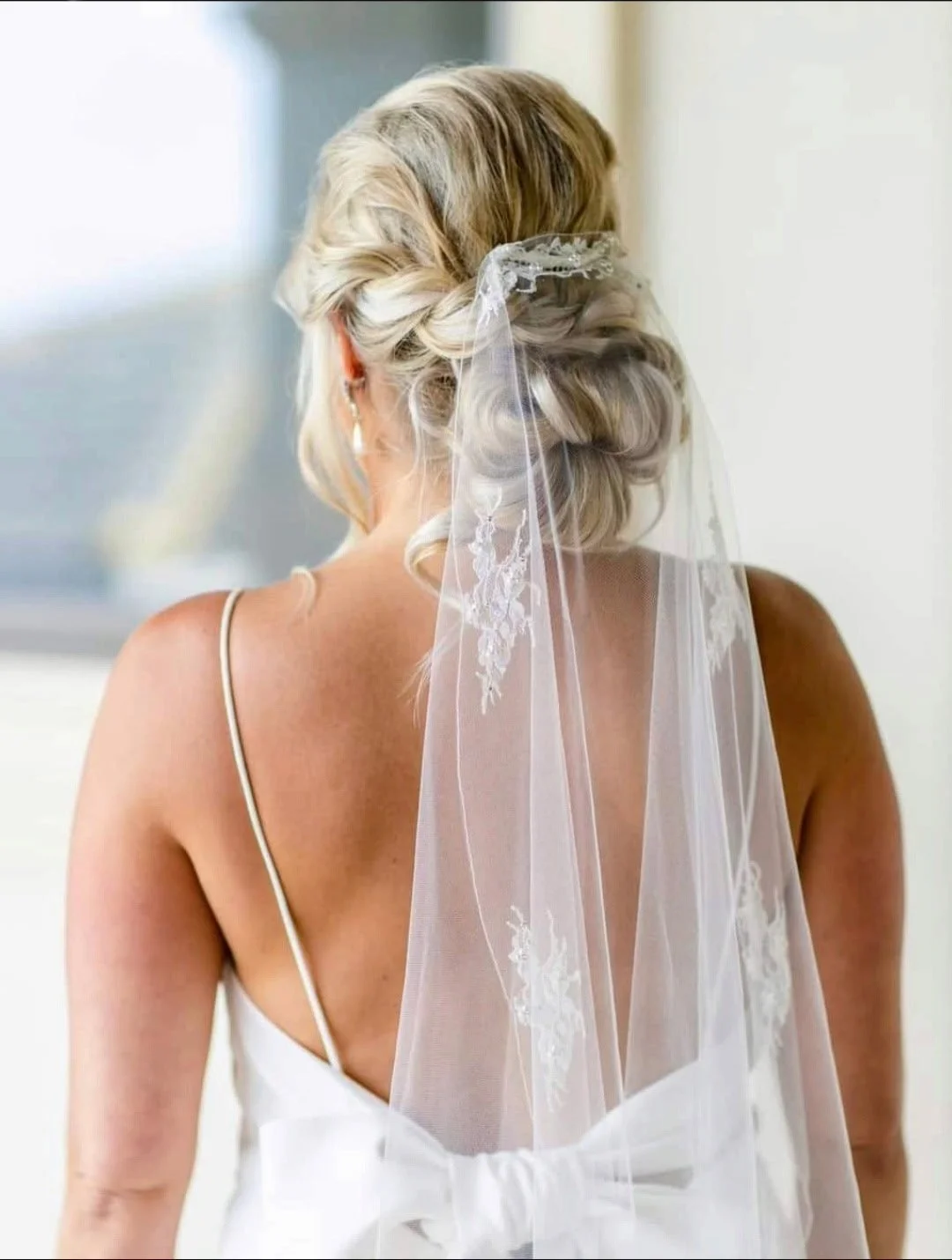 Back view of a bride with blonde hair styled in an elegant updo, wearing a white wedding dress with thin straps and a sheer veil with floral embroidery.