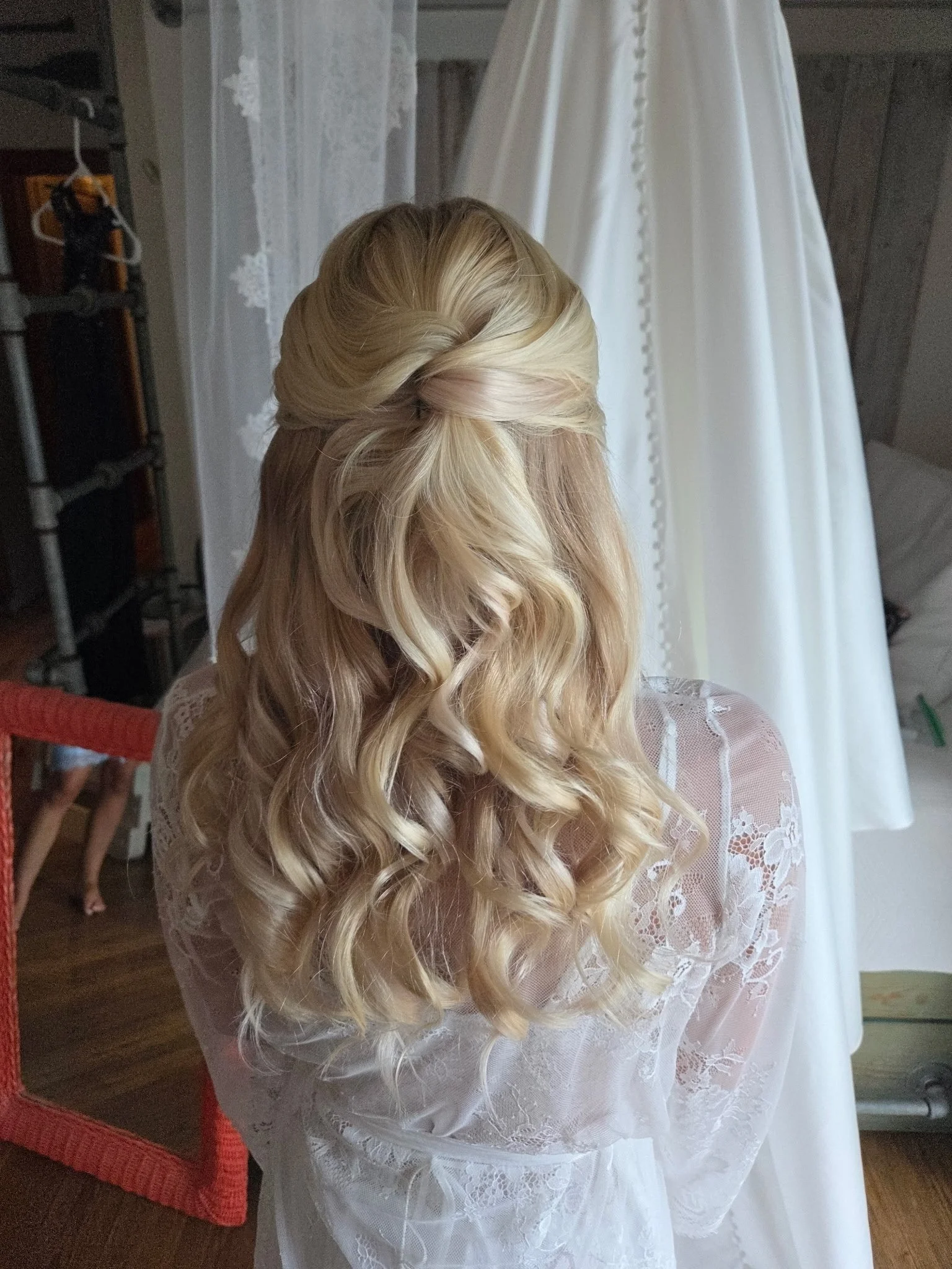 Woman with long, wavy blonde hair styled in an elegant half-updo, wearing a white lace garment, standing indoors near a white curtain and a wedding dress on a mannequin.