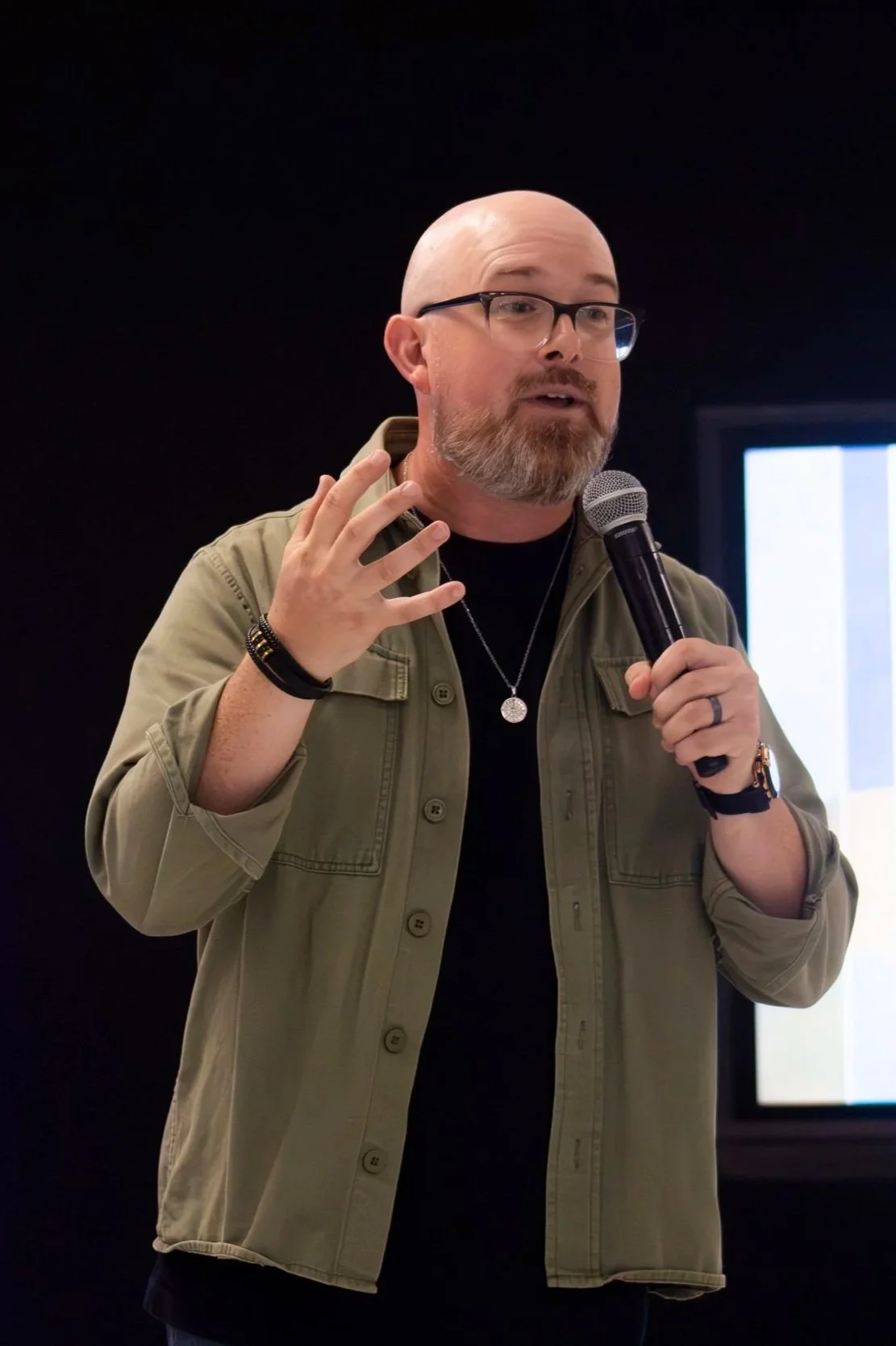 A man with glasses and a beard holding a microphone, speaking, wearing a green jacket, black shirt, and accessories, standing against a dark background.