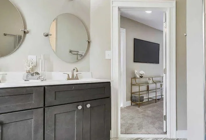 Bathroom with a double vanity with two round mirrors, a faucet, and glass containers on the counter, leading to a bedroom with a wall-mounted TV and a gold-colored console table.