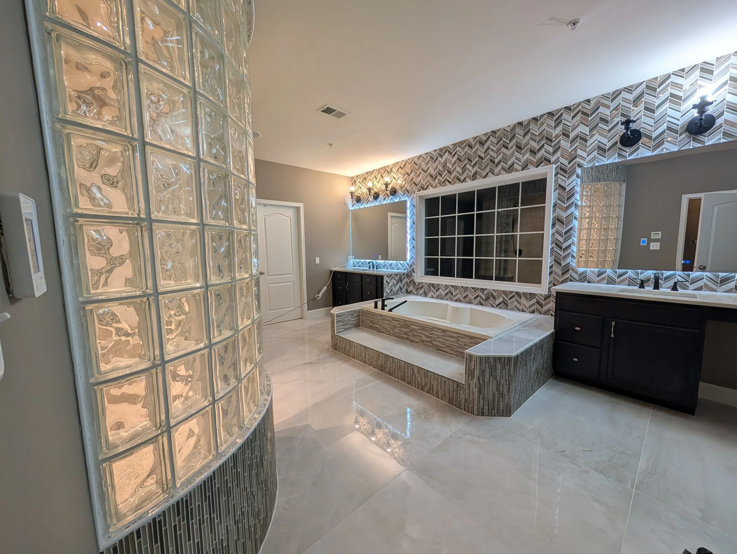 Modern bathroom featuring a built-in bathtub, black cabinetry, a large window, decorative tile walls, glass block wall, and shiny tiled floor.