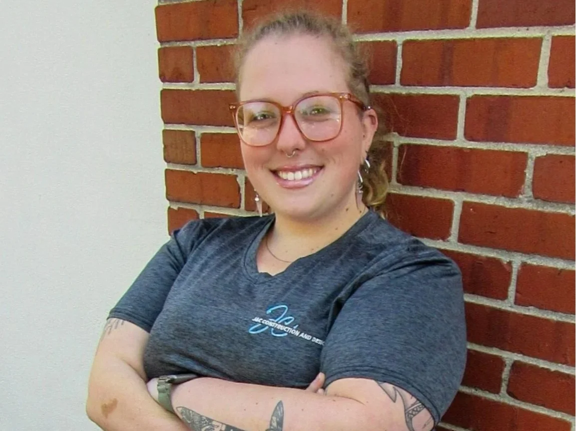 A woman with glasses and tattoos standing with her arms crossed, smiling, in front of a brick wall.