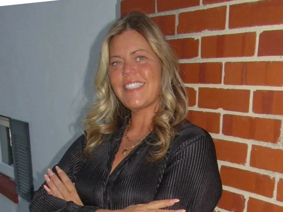 A woman with blonde hair, smiling, wearing a black long-sleeve shirt and matching pants, standing with arms crossed in front of a brick wall outside a building.