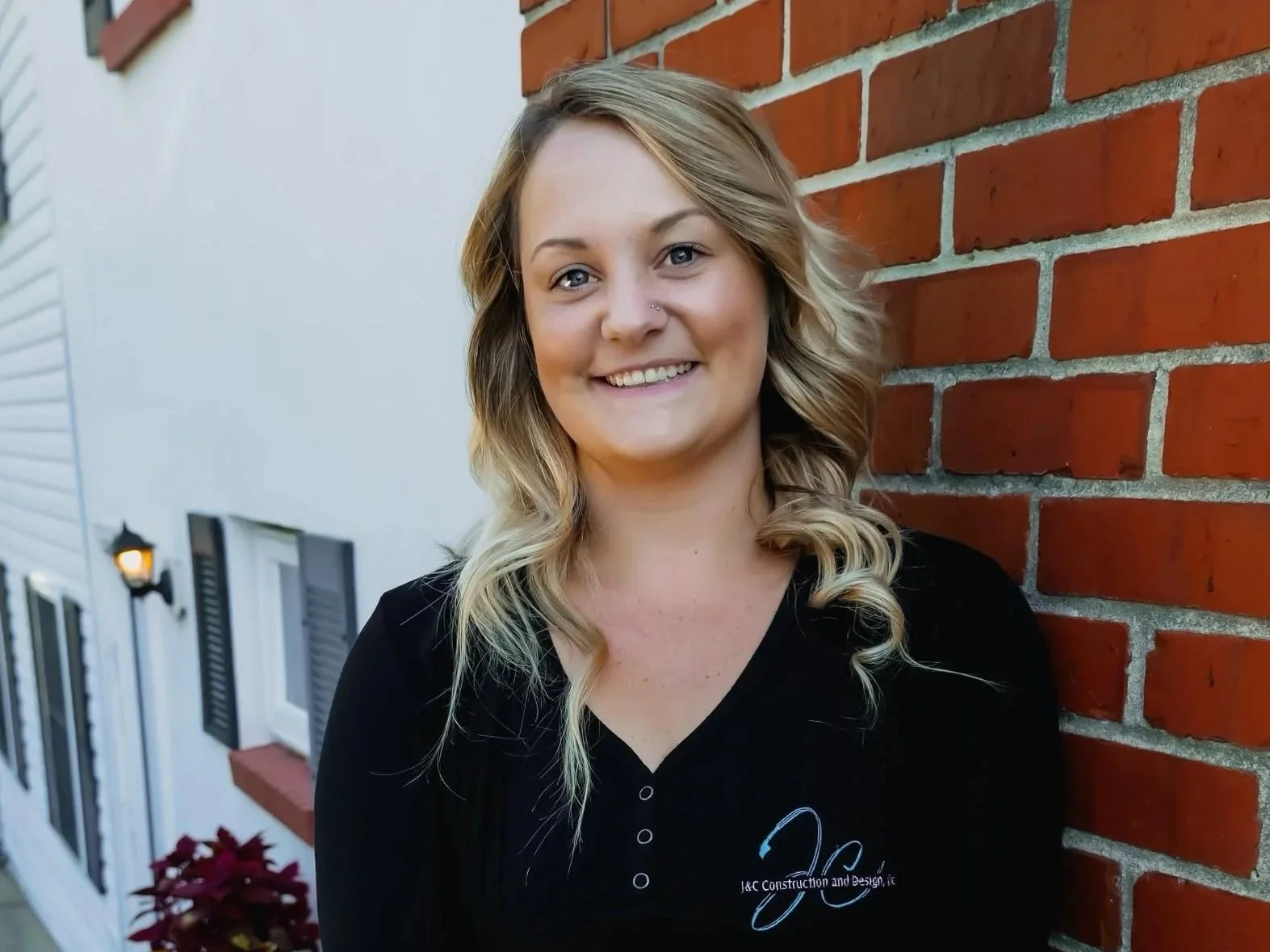 A smiling woman with wavy blonde hair standing outdoors, leaning against a brick wall. She is wearing a black shirt with embroidery and is crossing her arms. In the background, there is a white house with black shutters, a small potted plant with red leaves, and a lawn.