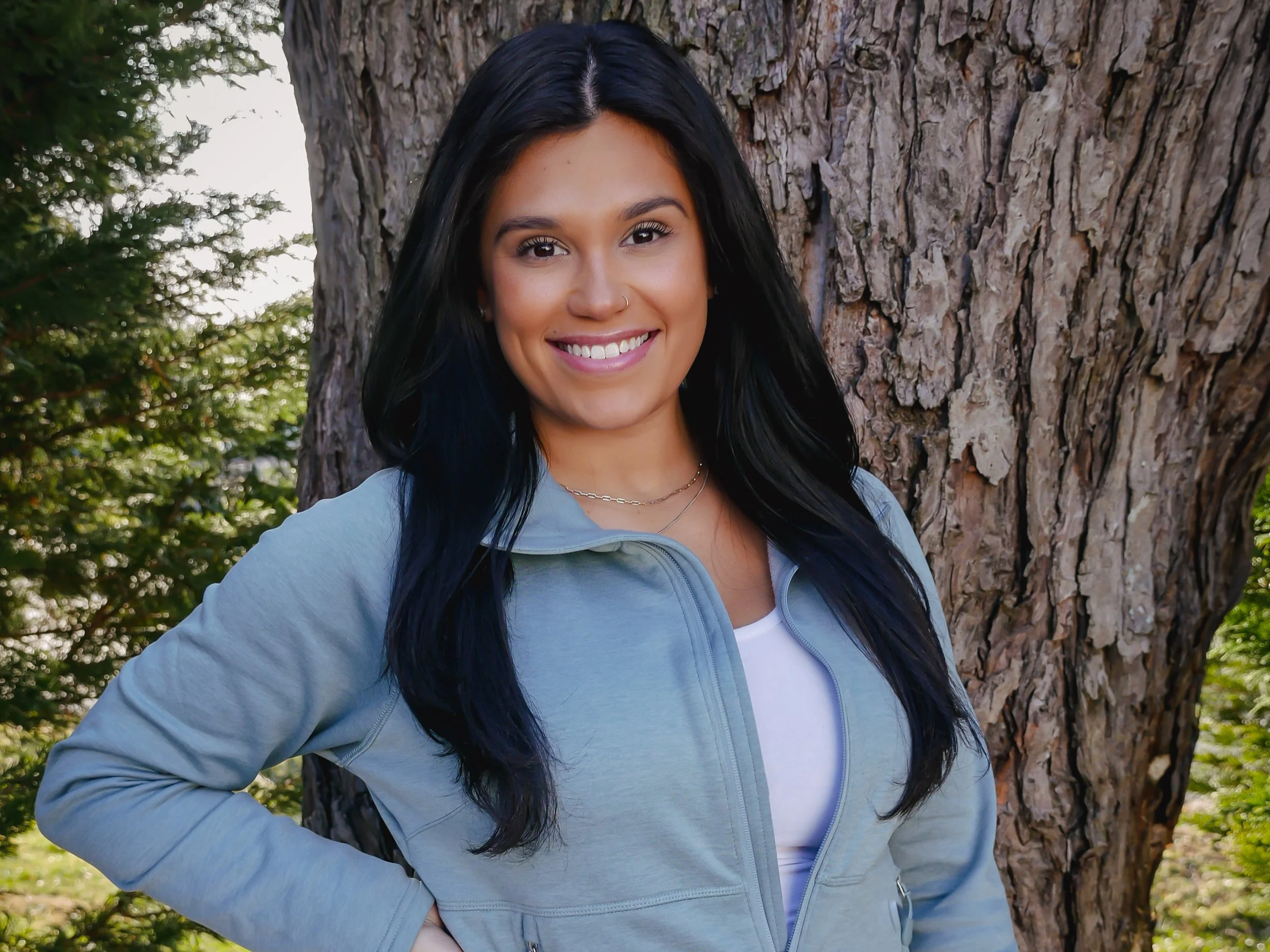 A woman with long black hair, wearing a light blue zip-up jacket over a white shirt, standing outdoors in front of a large tree with green foliage in the background, smiling at the camera.