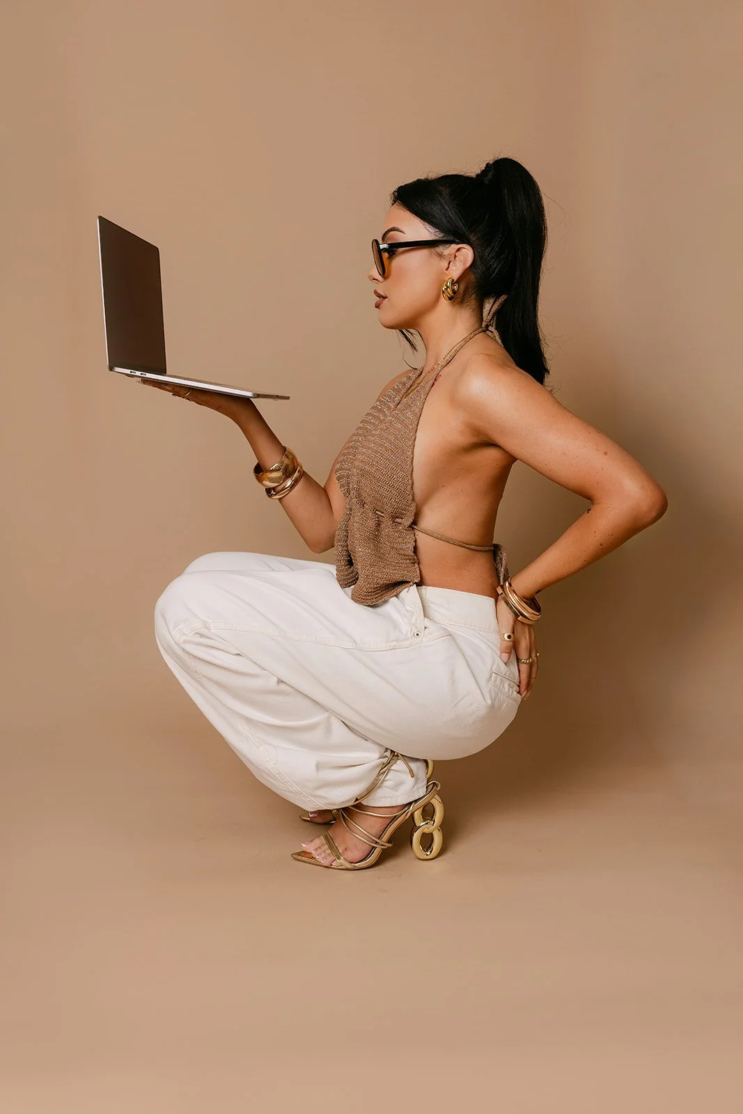 A woman crouching on the floor, holding a laptop in her right hand, wearing sunglasses, gold jewelry, a backless top, white pants, and high-heeled sandals with a chain detail, against a beige background.