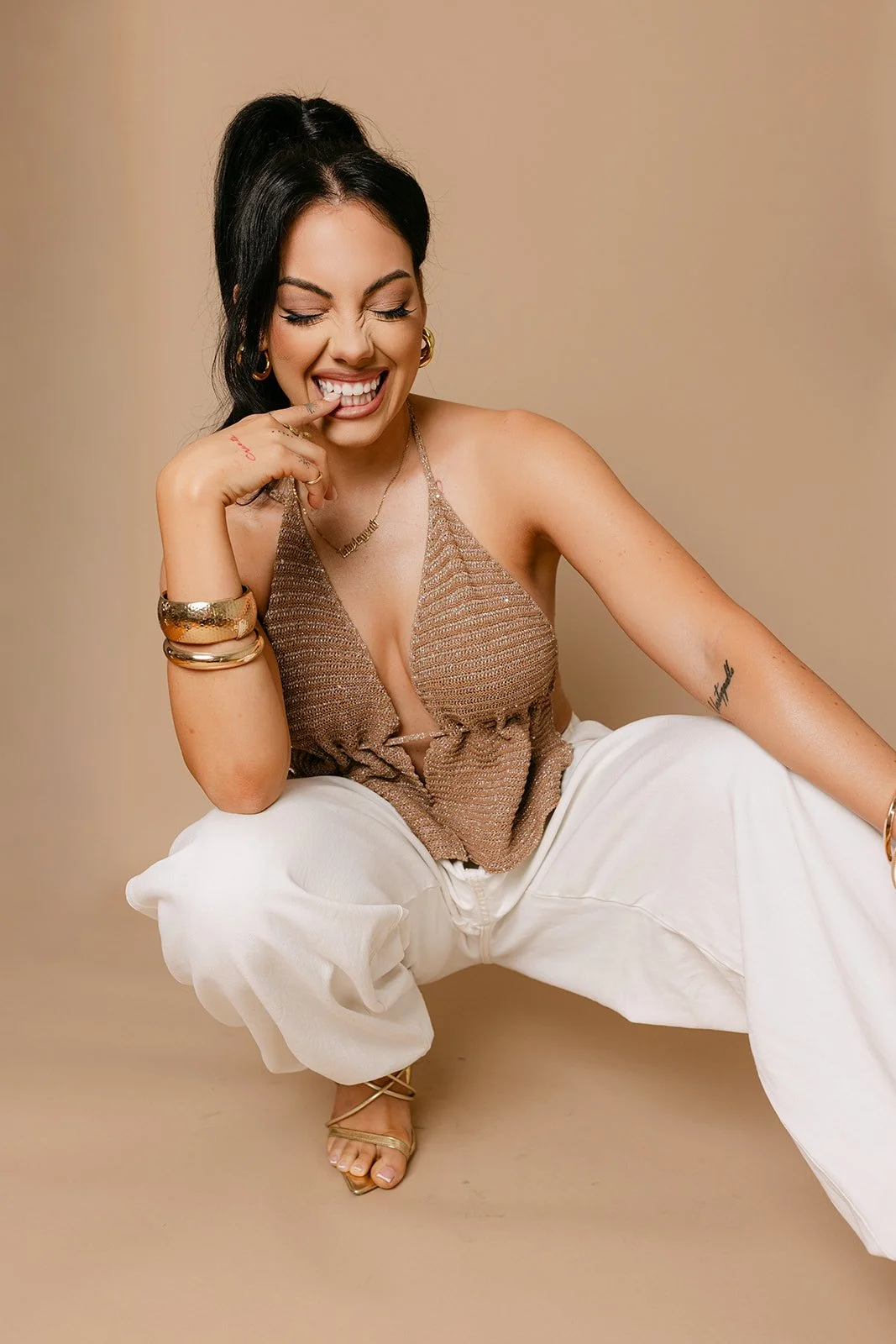 A woman with dark hair tied up in a ponytail, wearing gold jewelry, a beige sleeveless top, and white pants, squatting on the floor and smiling playfully while touching her lips with her finger, against a beige background.