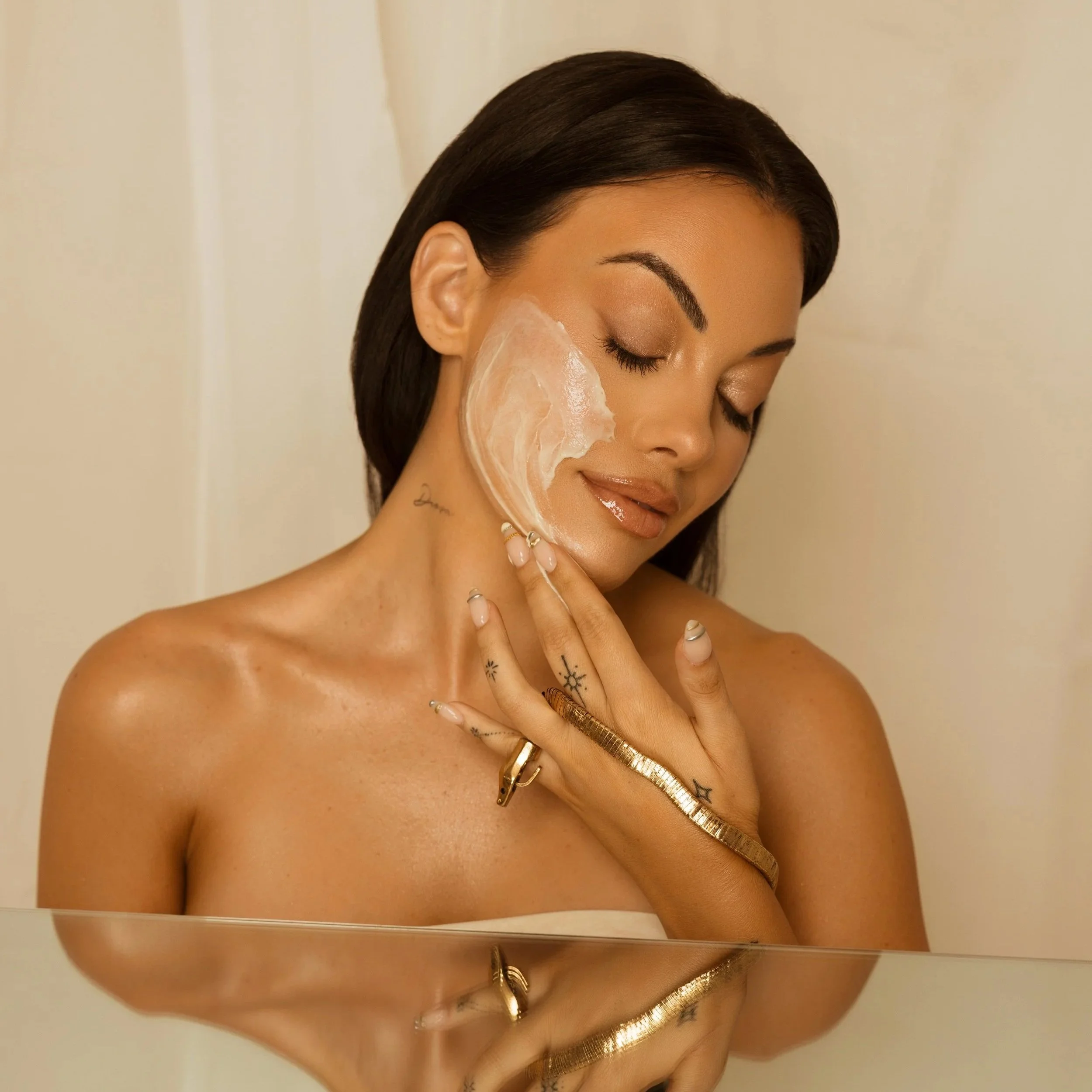 A woman with dark hair and tattoos on her hand and fingers, applying a cream or lotion to her face with her eyes closed against a beige background.