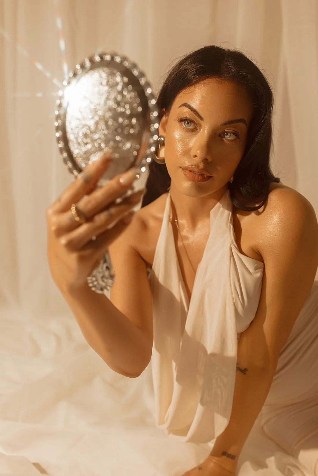 A woman with dark hair and light-colored eyes taking a selfie in a mirror, holding a decorative silver handheld mirror, with her face and upper body visible, wearing a sleeveless, light-colored top with a bow tie, and jewelry including earrings and rings.