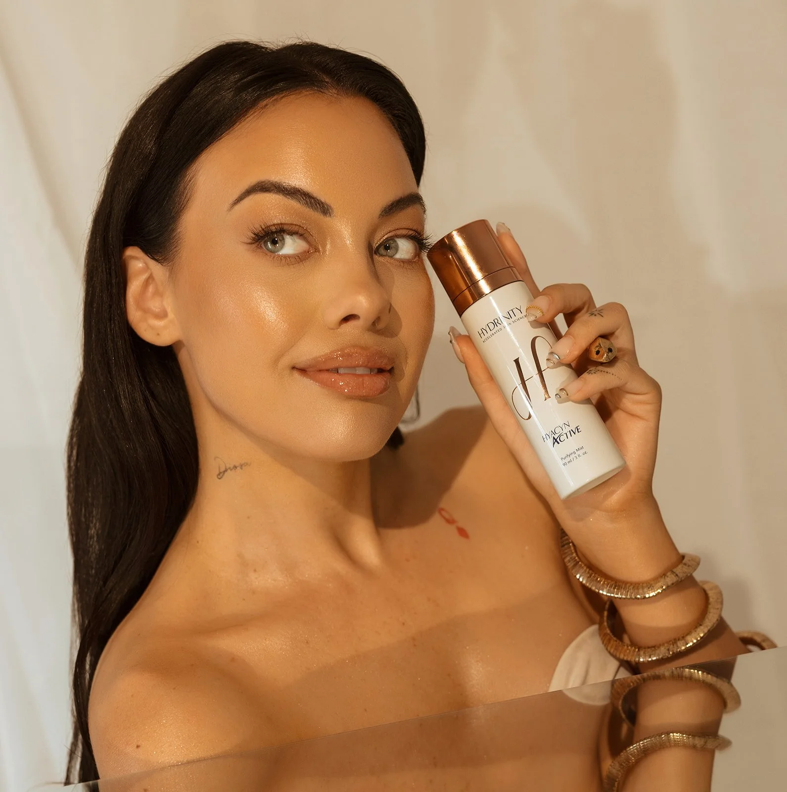 A woman with long dark hair holding a white and copper bottle of HydraVity Hyacinth Active skincare spray, smiling with a glowing complexion, wearing multiple bracelets on her wrist.