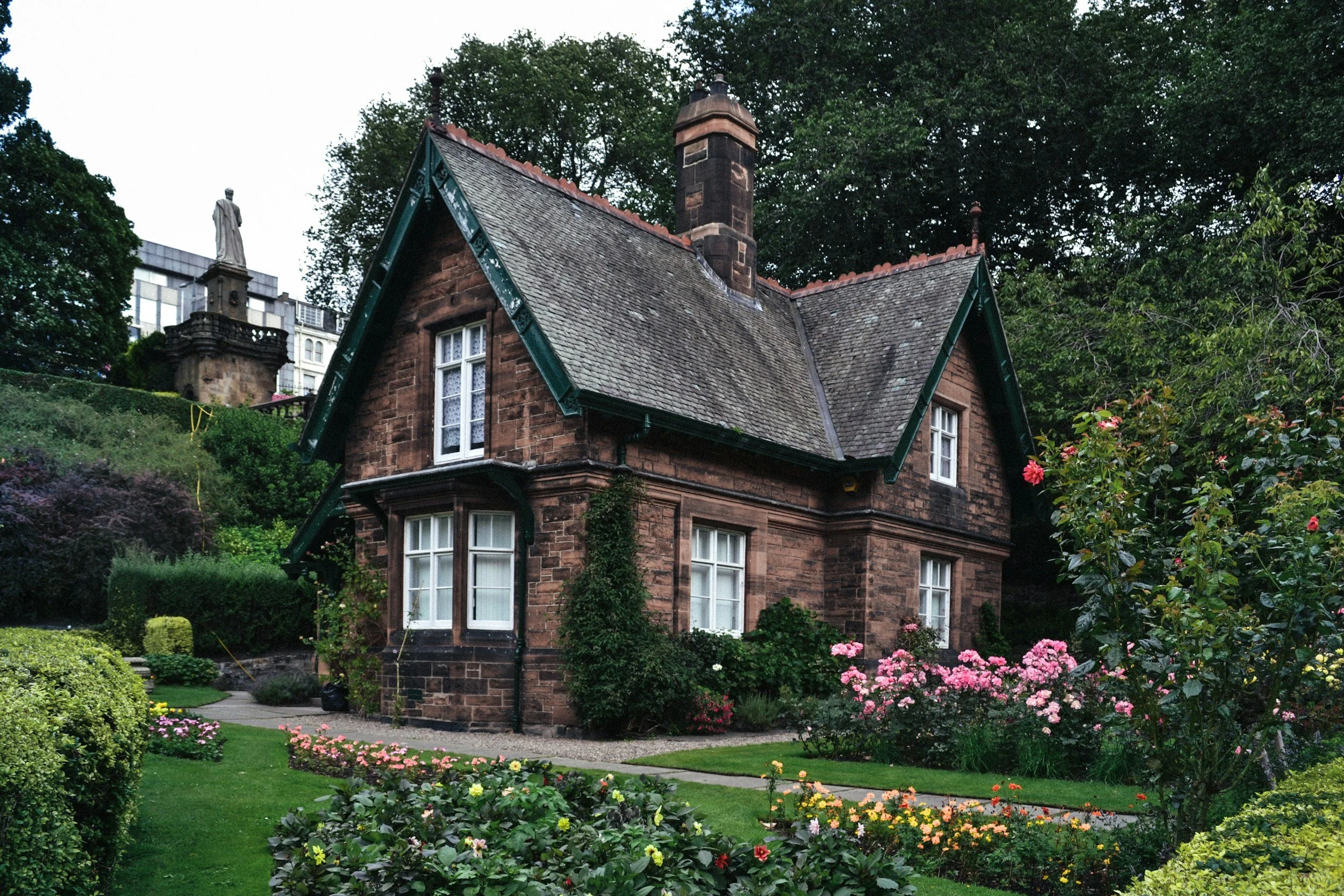 A brick house with a steep roof, situated in a lush garden with colorful flowers and trimmed bushes. There are tall trees and a statue on a pedestal in the background.