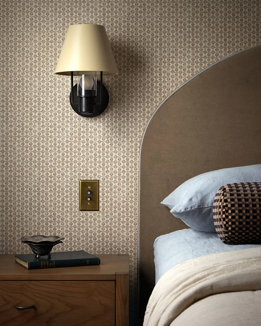 Brass sconce and a curved upholstered headboard with patterned wallpaper in a Vestige Home Philadelphia bedroom